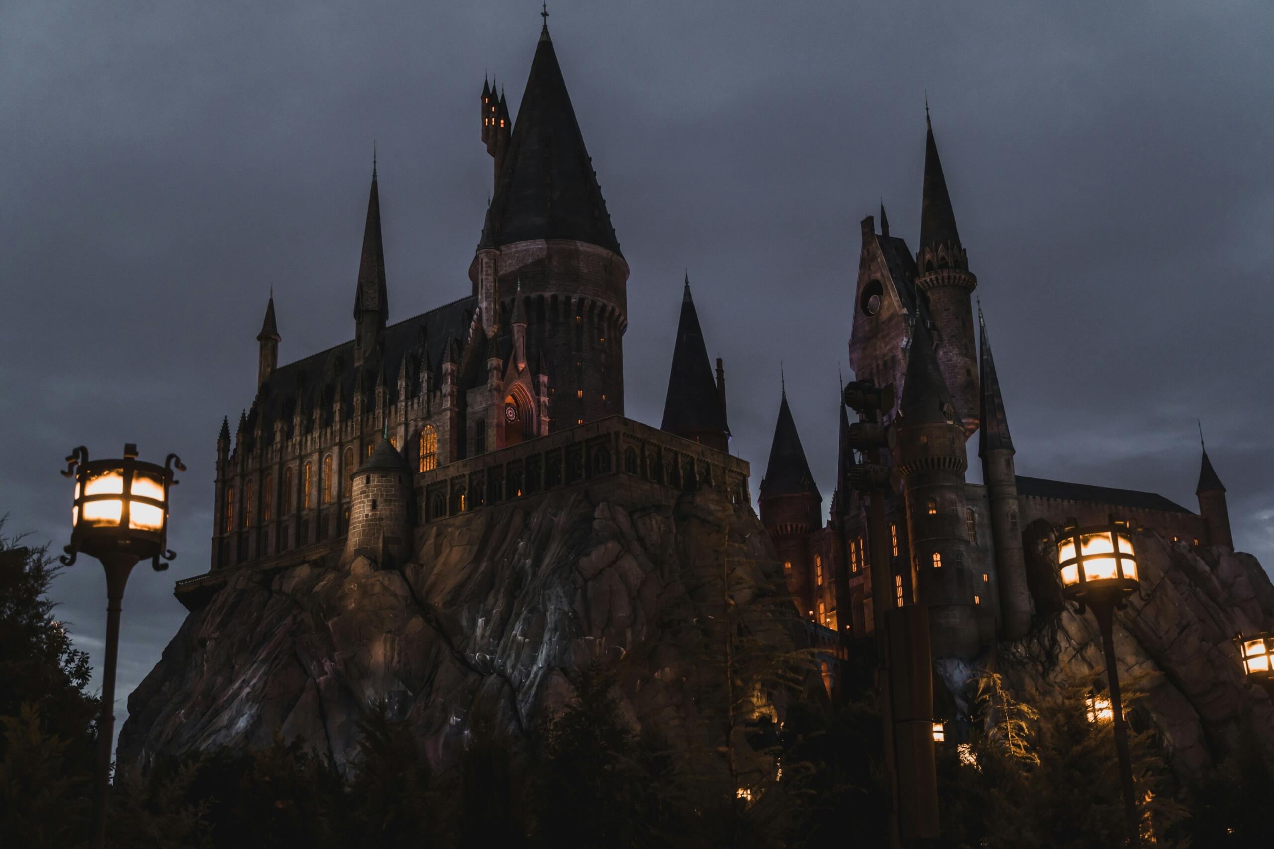 Veliki kameni dvorac Hogwarts s visokim tornjevima na stijeni, osvijetljen toplim svjetlima u sumrak pod oblačnim nebom.