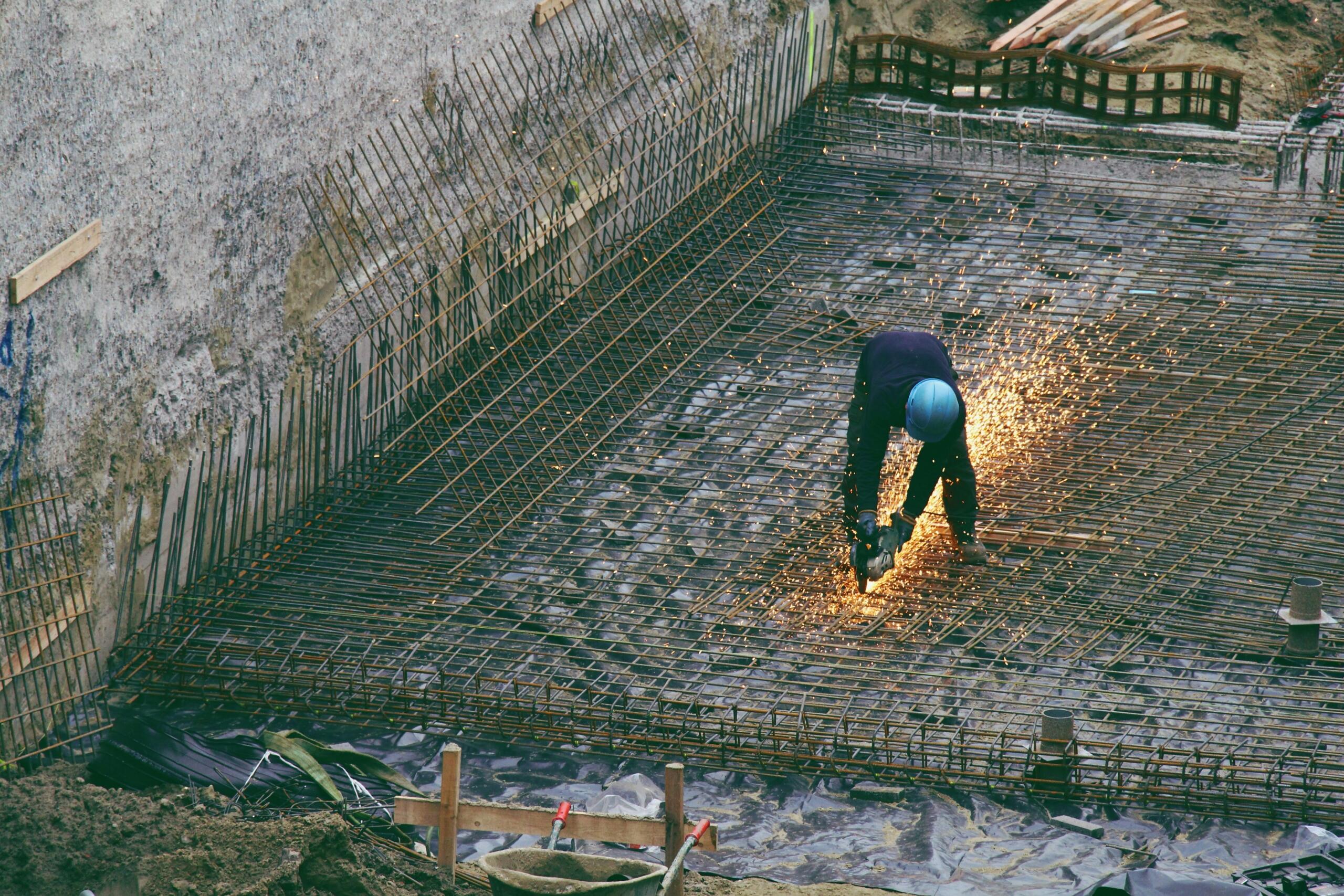 Homonimi na engleskom – radnik sa plavim zaštitnim šljemom reže metalnu armaturu na velikom građevinskom gradilištu (construction site), dok iskre pršte svuda oko njega.