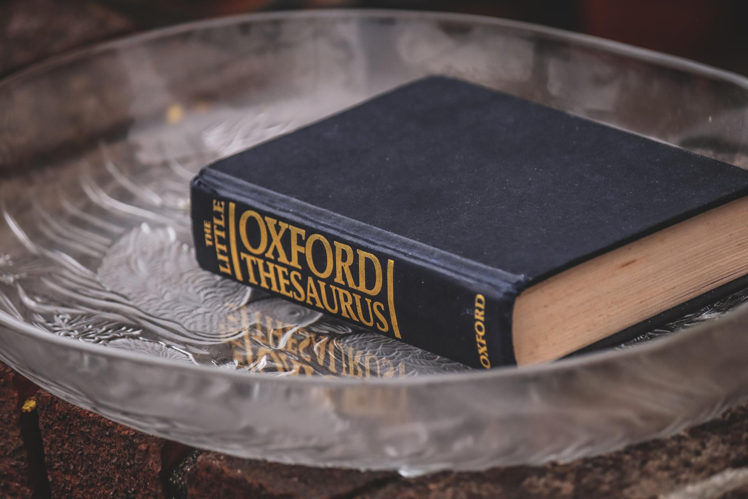 Kako se izgovara na engleskom – knjiga Oxford Thesaurus sa crnim koricama i zlatnim slovima položena u staklenu ukrasnu posudu.