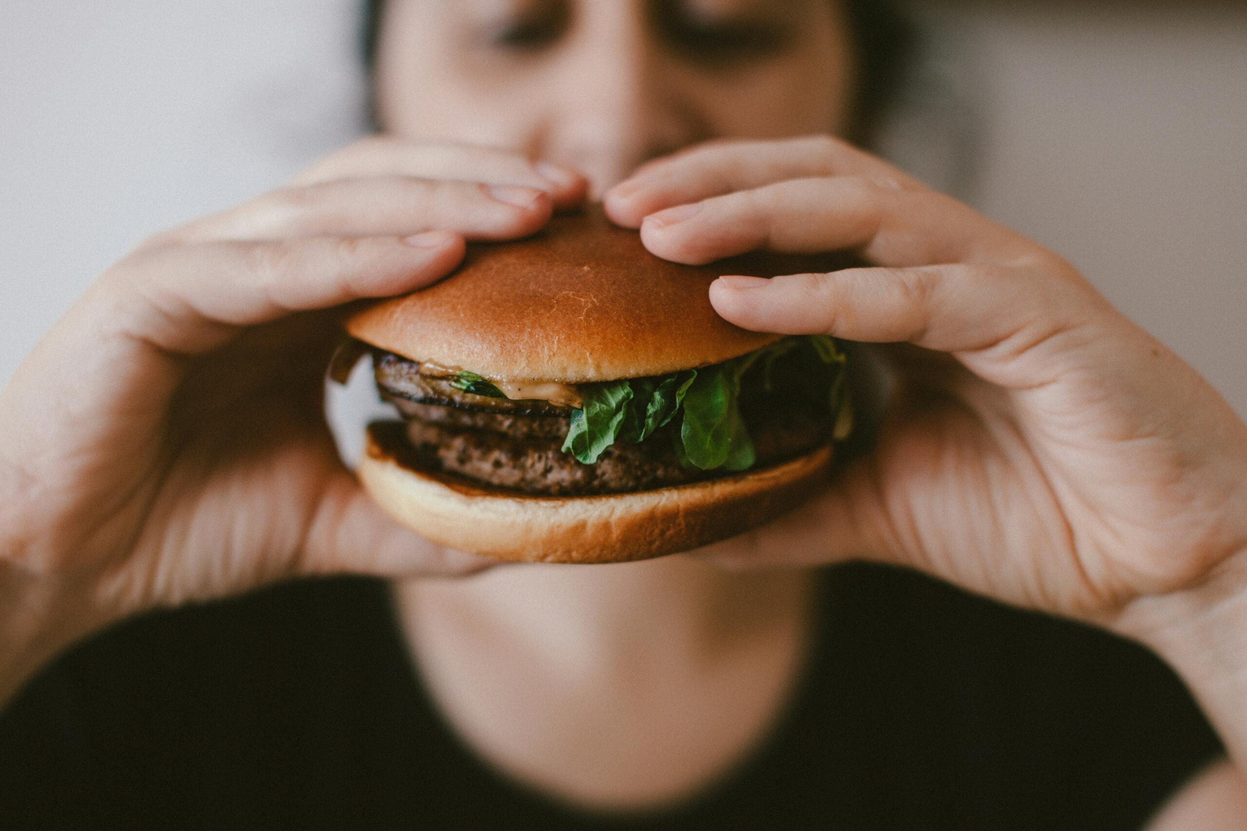 Zanimljive engleske riječi – krupni plan osobe koja objema rukama drži sočan, veliki hamburger sa zelenom salatom i prinosi ga ustima.