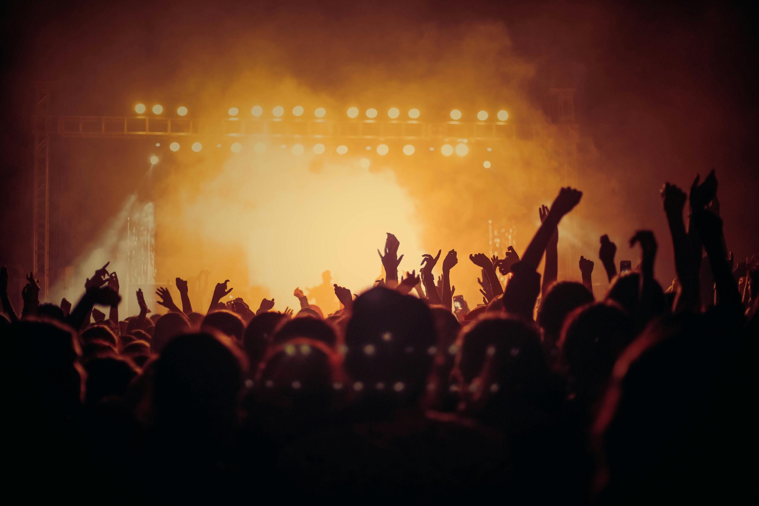 Publika na muzičkom koncertu s podignutim rukama ispred osvijetljene bine pod toplim žutim svjetlima