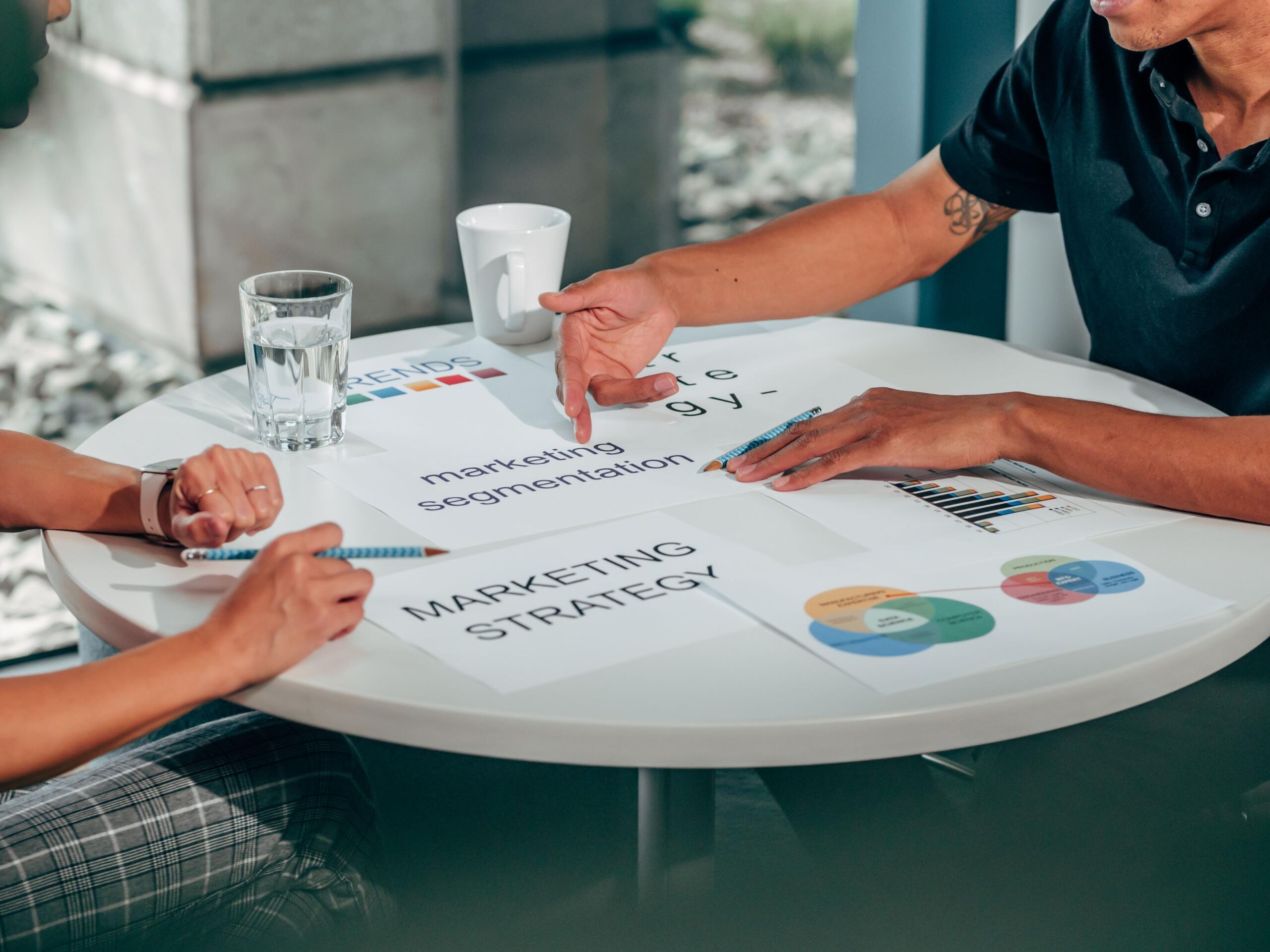 Dvije osobe sjede za okruglim stolom i pokazuju na dokumente sa natpisima Marketing Strategy i Marketing Segmentation
