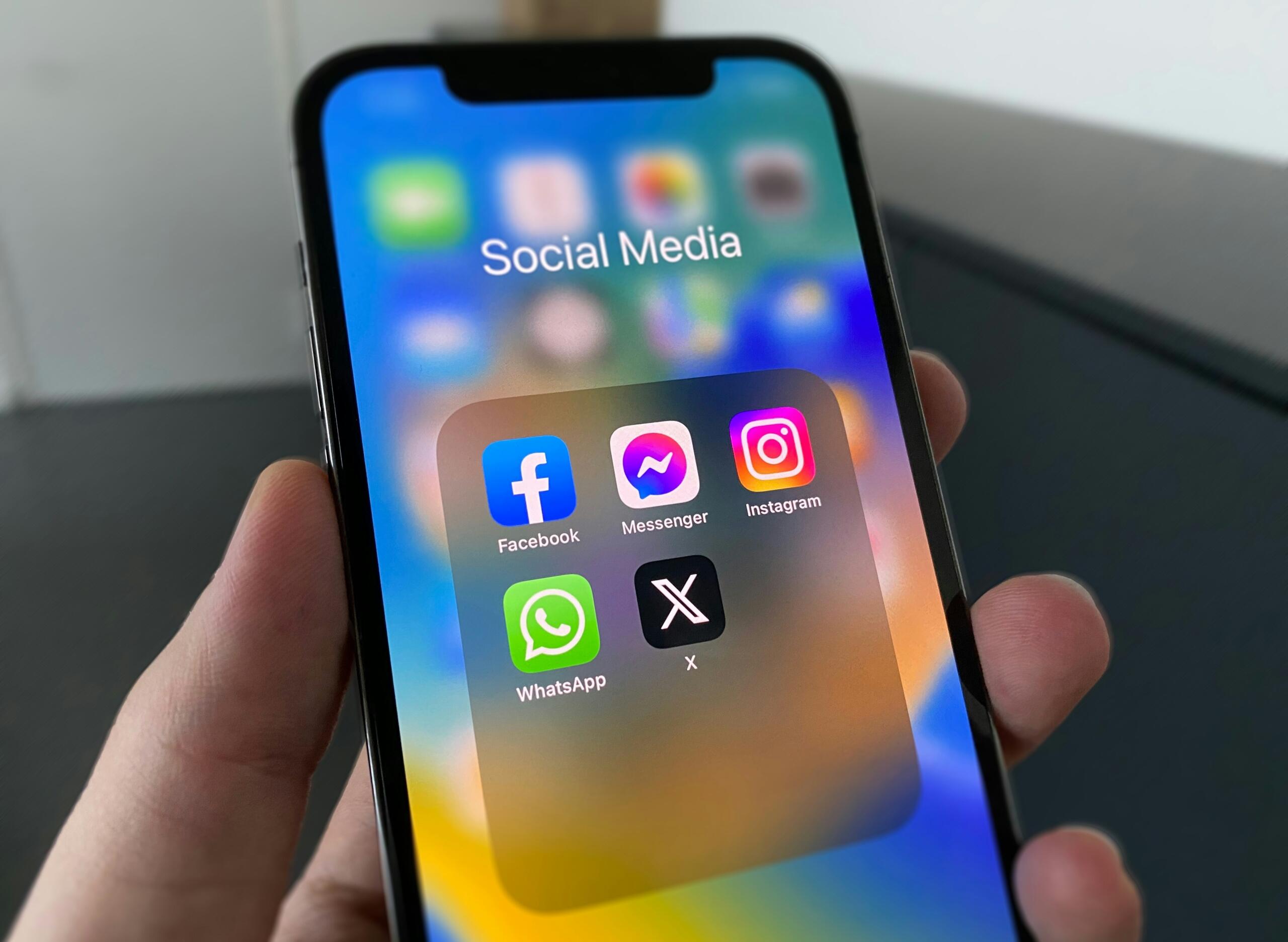 Pametni telefon u ruci sa prikazanim folderom aplikacija za društvene mreže (Facebook, Instagram, Messenger, WhatsApp, X)