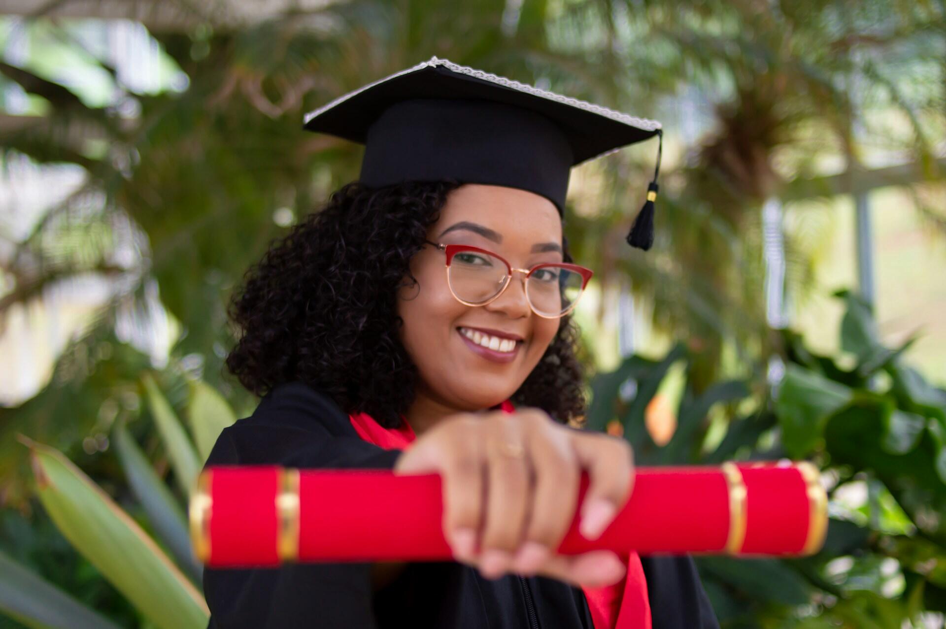 jeune femme aux cheveux noirs boucles et a lunette en toje tenant son diplome rouge