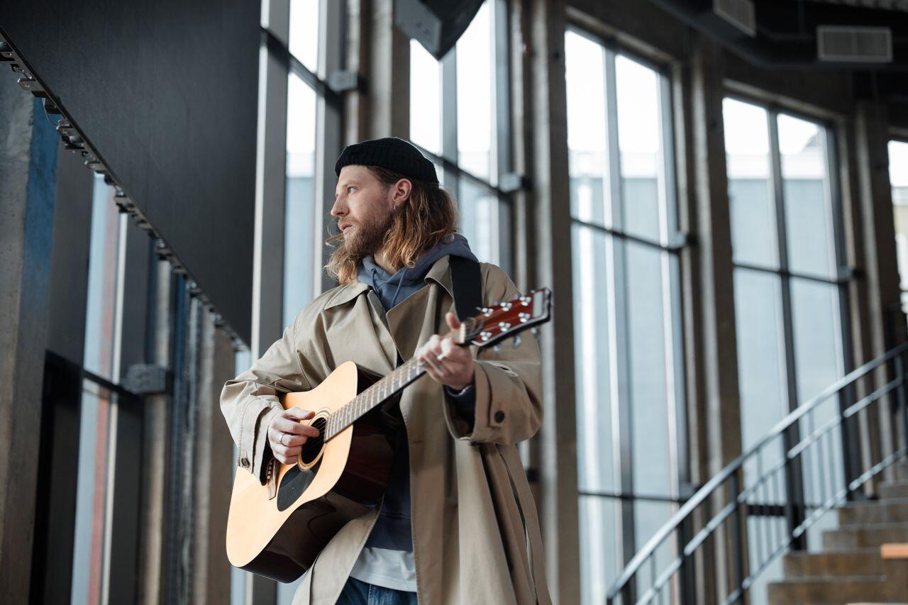 homme avec beret et cheveux longs avec la guitare devant des baies vitrees