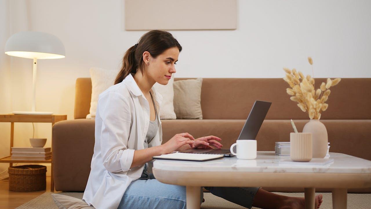 jeune femme en jean et chemise blanche assise par terre devant la table basse du salon avec son ordi et un cafe