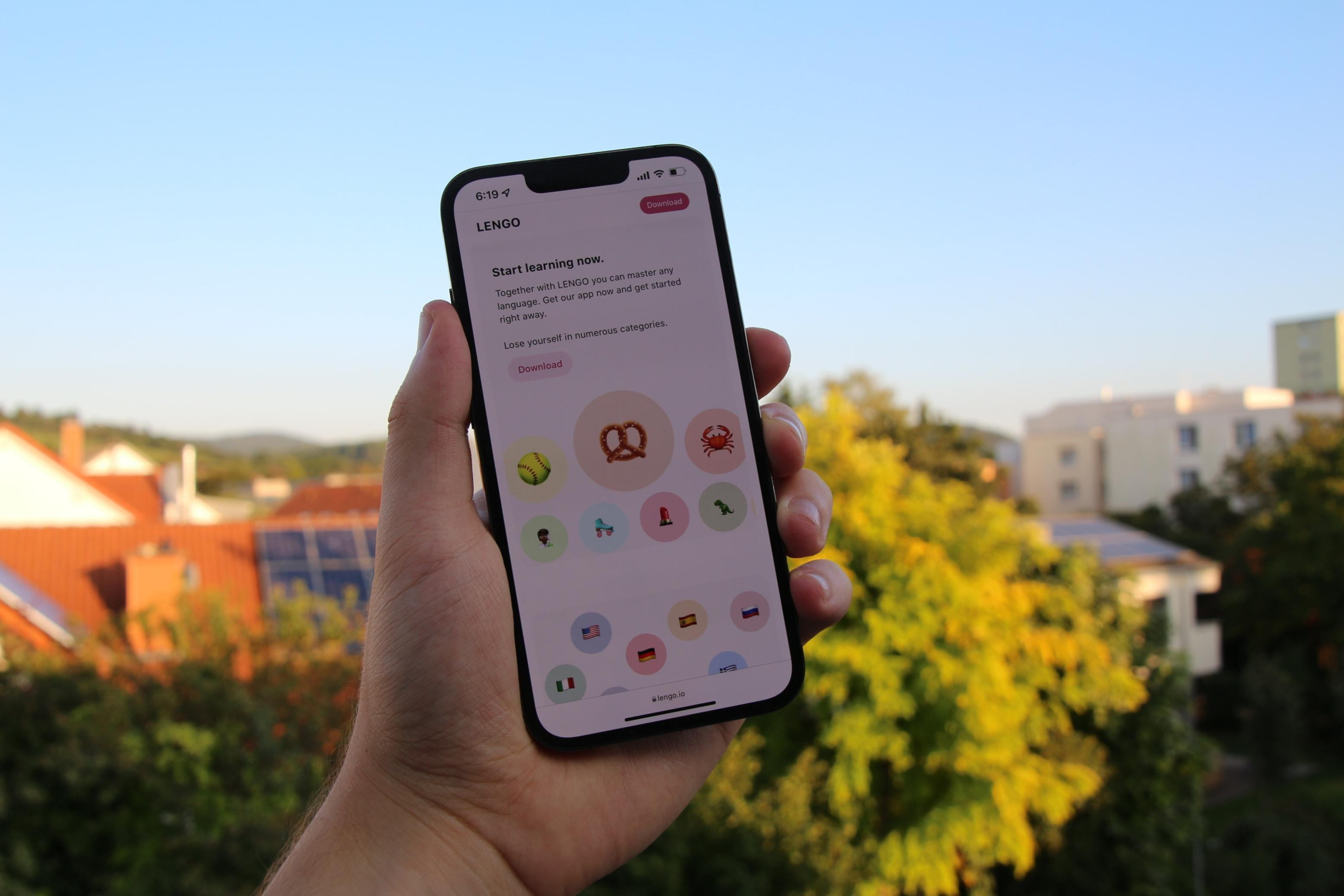 Une main tient en un téléphone qui montre une application qui permet d'apprendre une langue étrangère. Fond avec des arbres et des maisons.