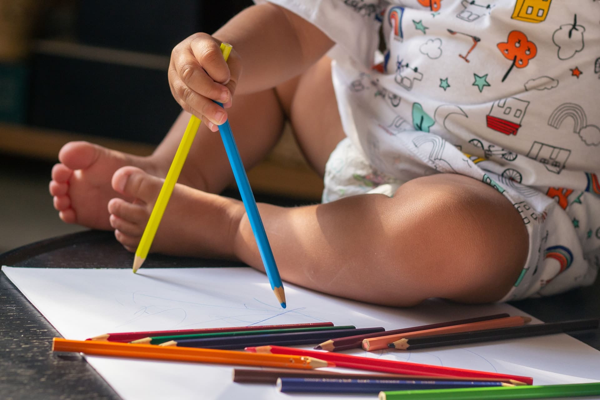 Bebê sentado no chão desenha co lápis de cor sobre papel.
