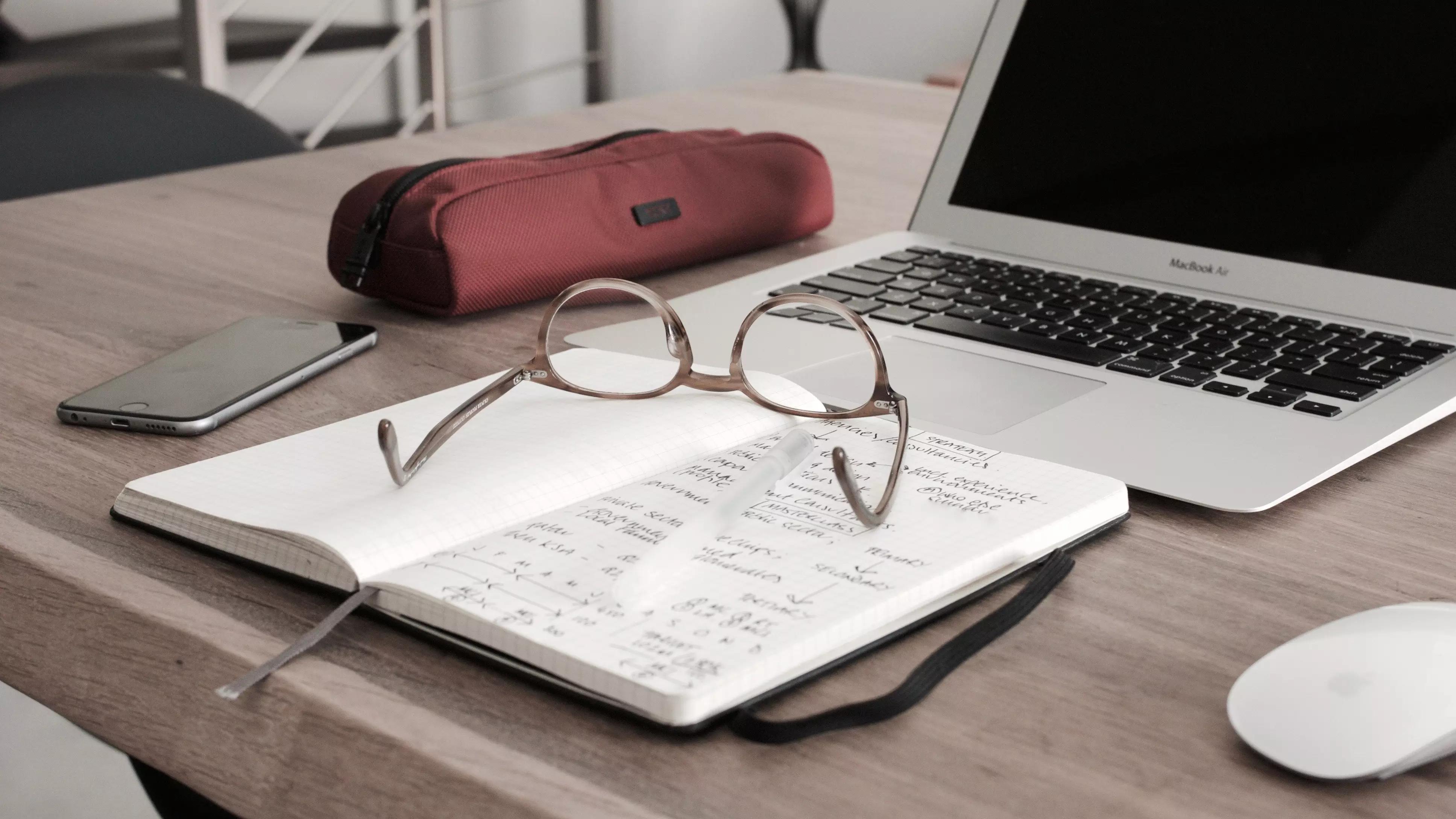 Um notebook, um caderno de notas e um óculos sobre uma mesa.