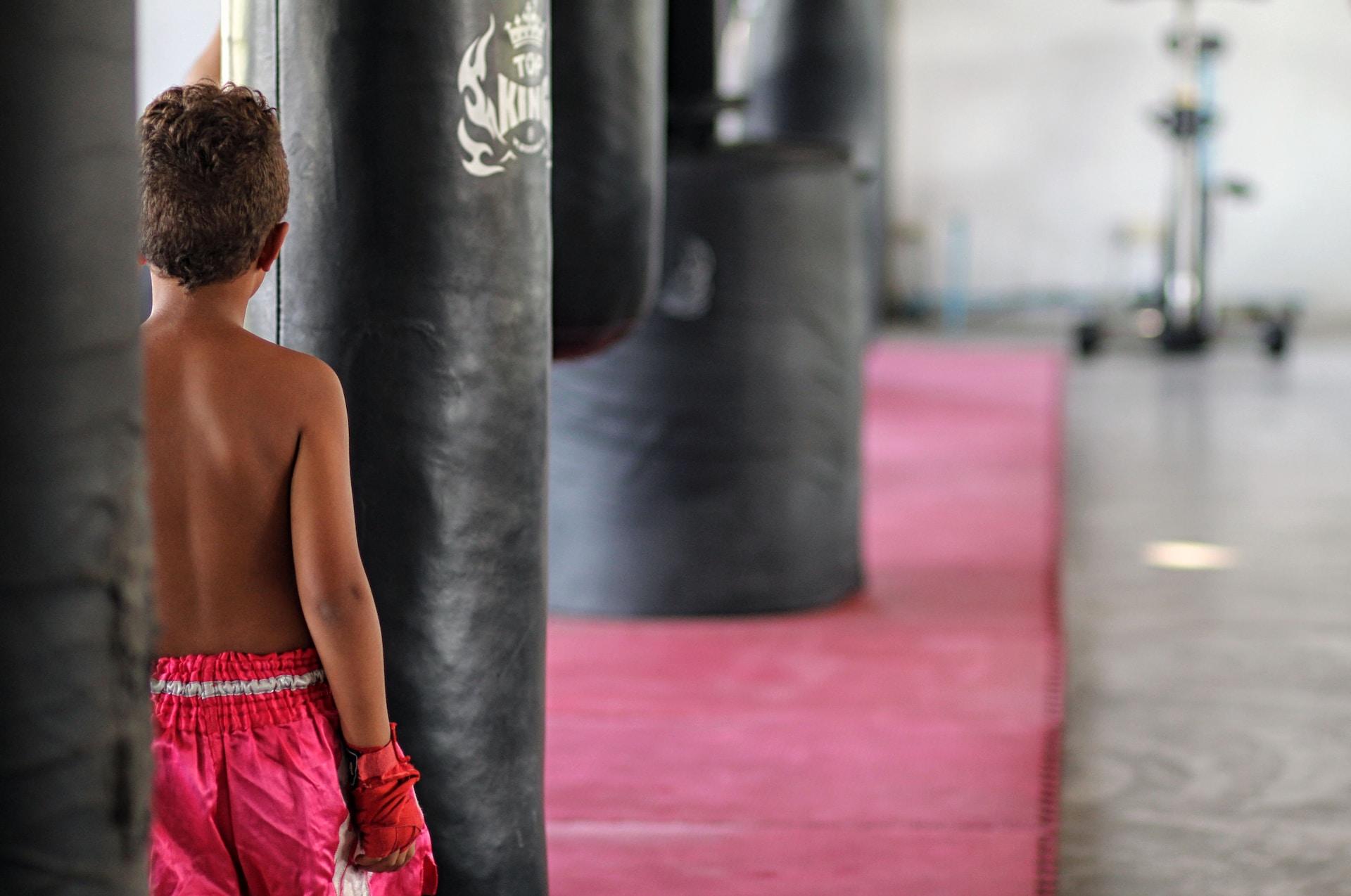 menino de costas em uma sala cheia de sacos de boxe, vestindo short e luvas de boxeador