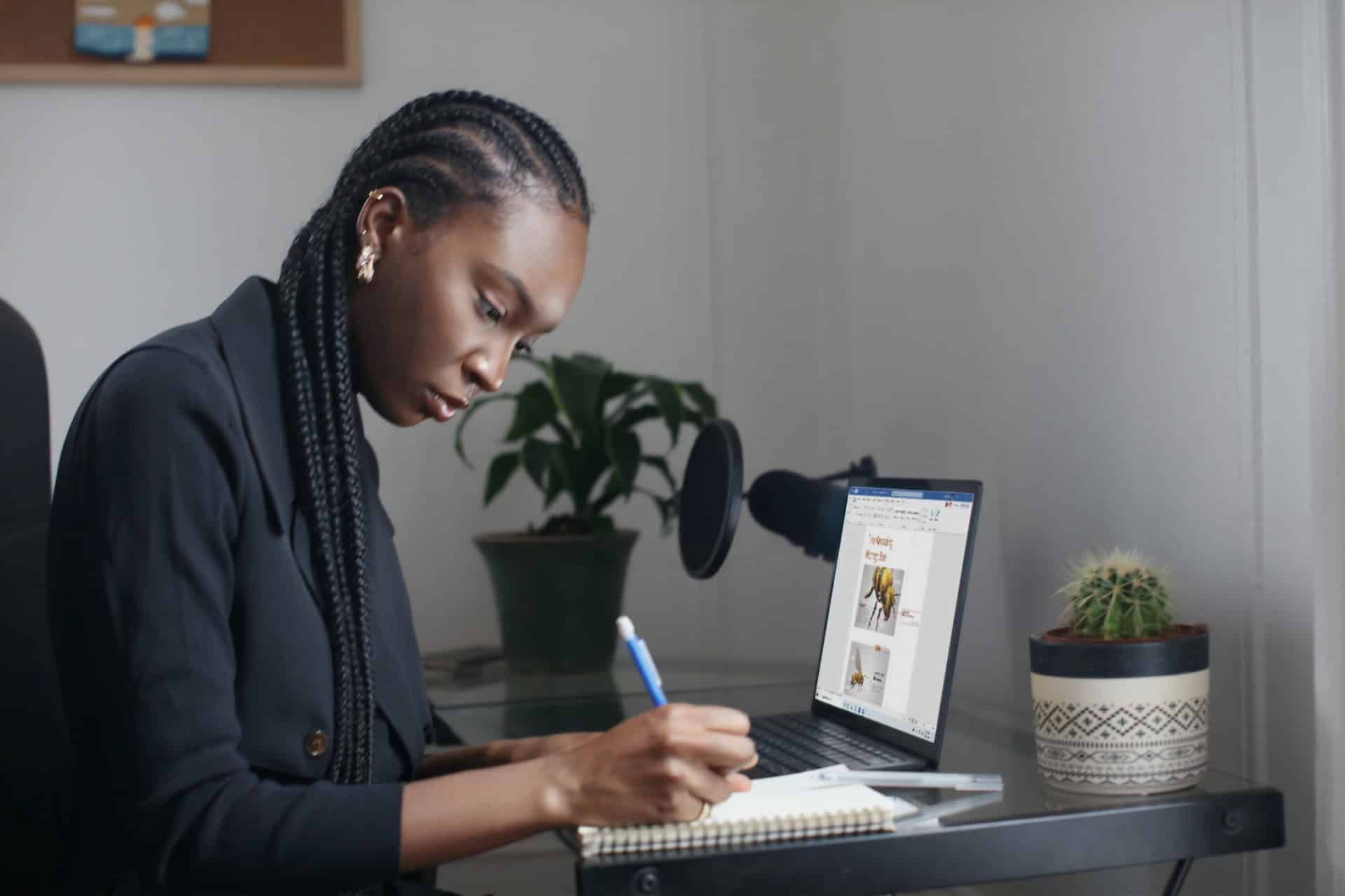 mulher negra, escrevendo em um caderno e com o pc ligado ao lado