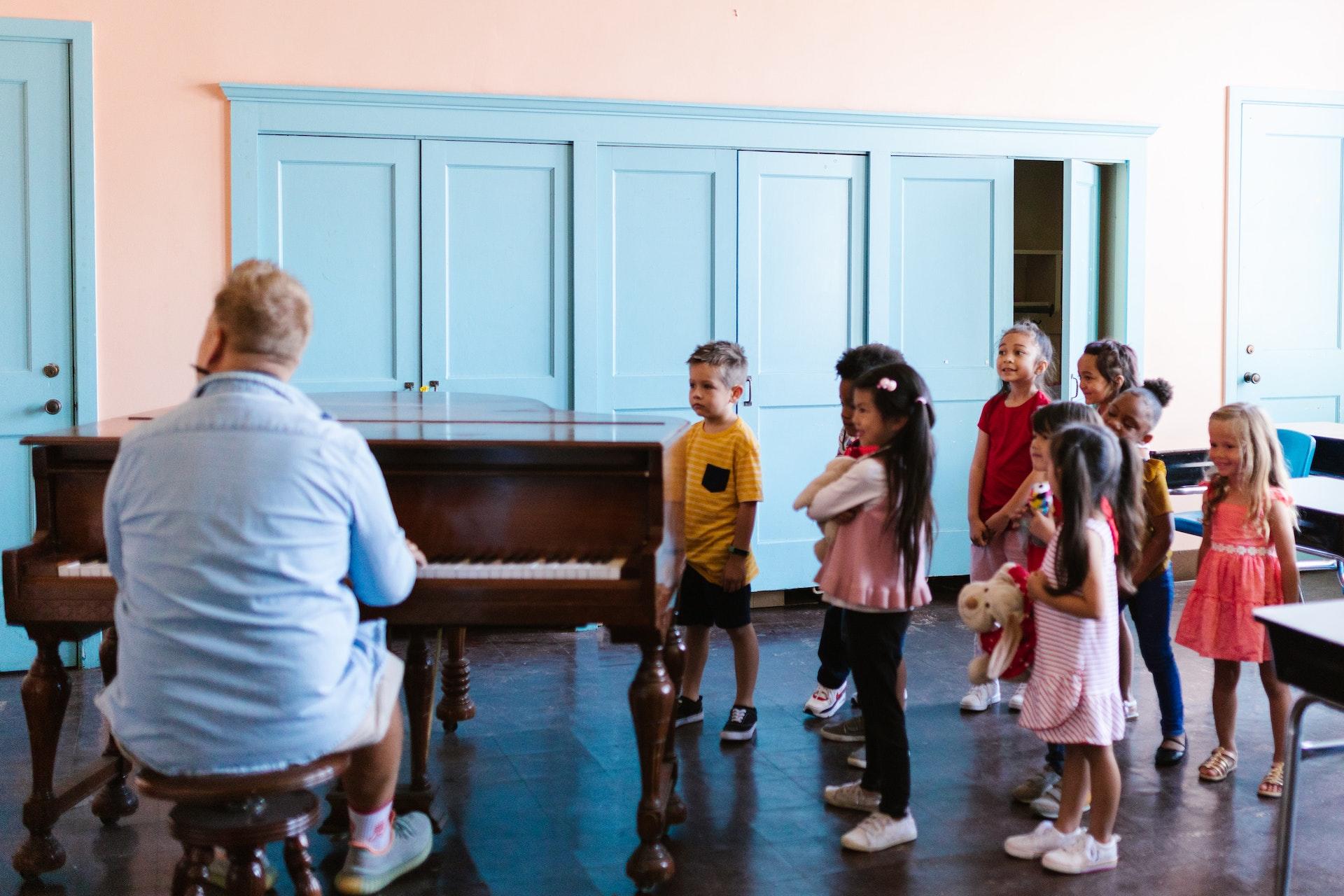 Sala com um grande piano e um professor tocando, com diversas crianças ao redor. Elas estão descontraídas e algumas trazem brinquedos nas mãos. 