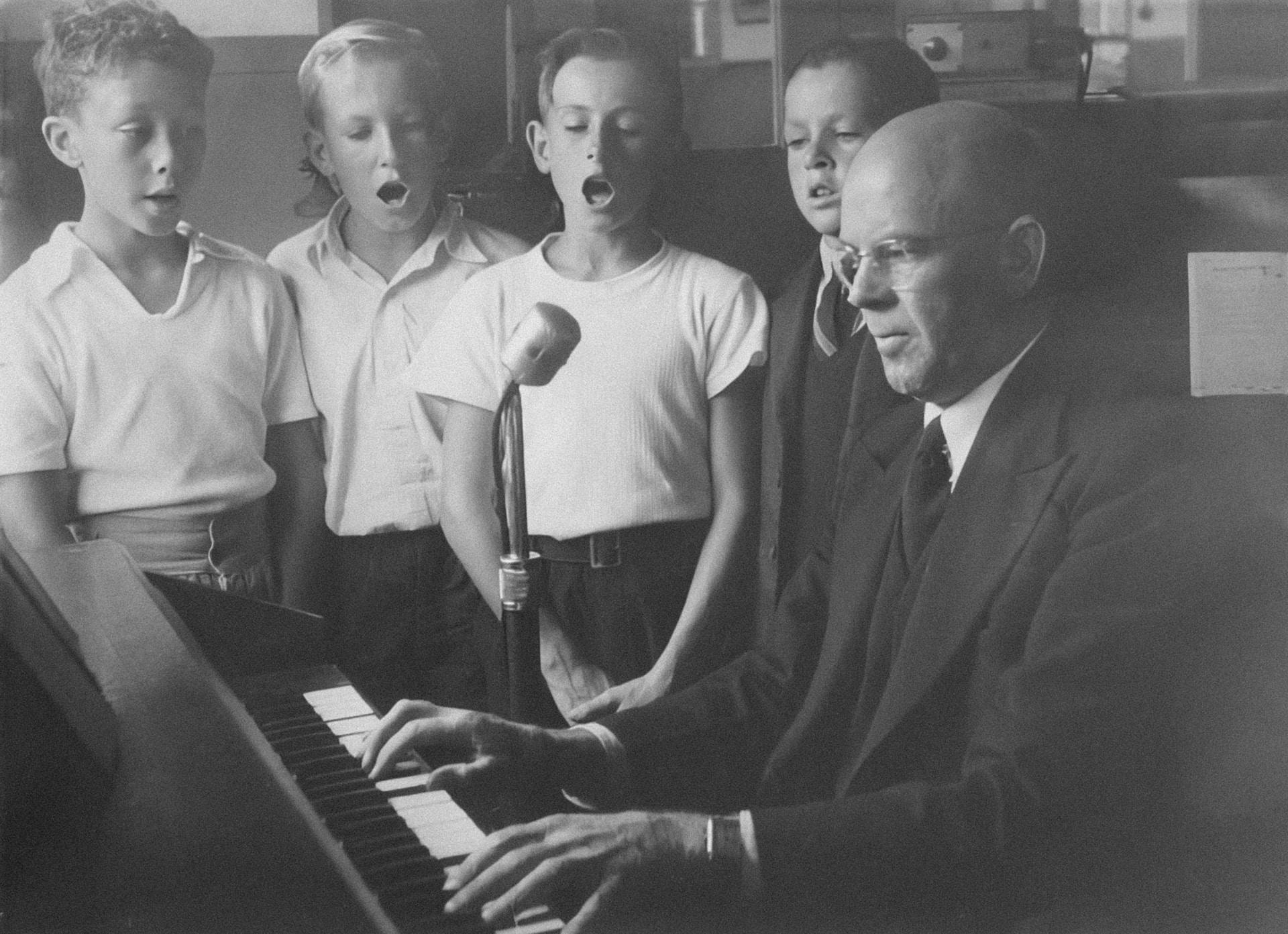professor dando aula de canto em um piano, com alunos cantando