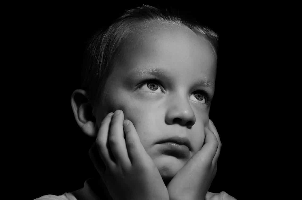 Menino com expressão triste apoia o queixo sobre as mãos e seu olhar infere que está divagando.