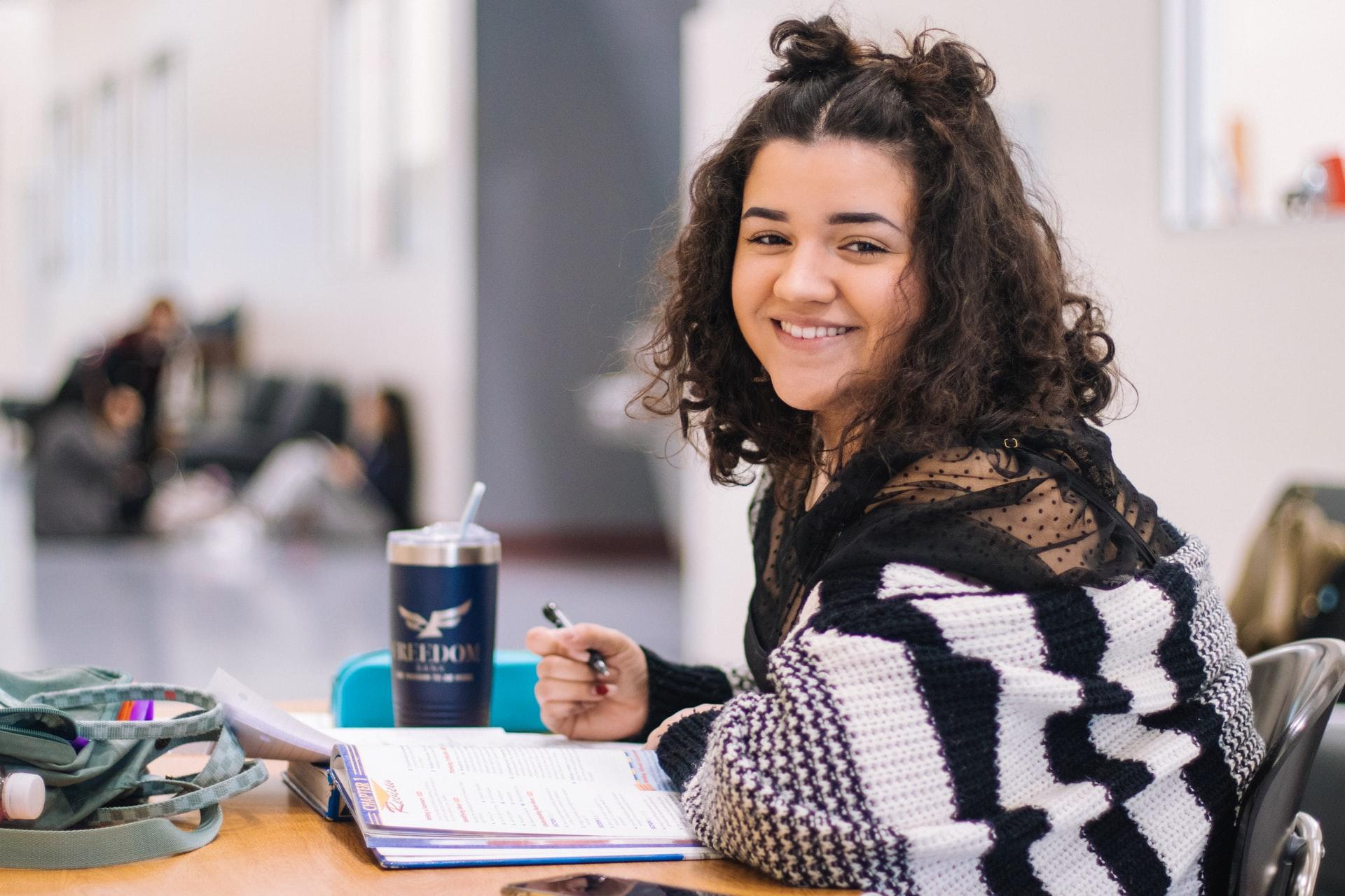 Jovem estudante está diante de uma mesa com o caderno aberta e, segurando um lápis, ela sorri para a câmera.
