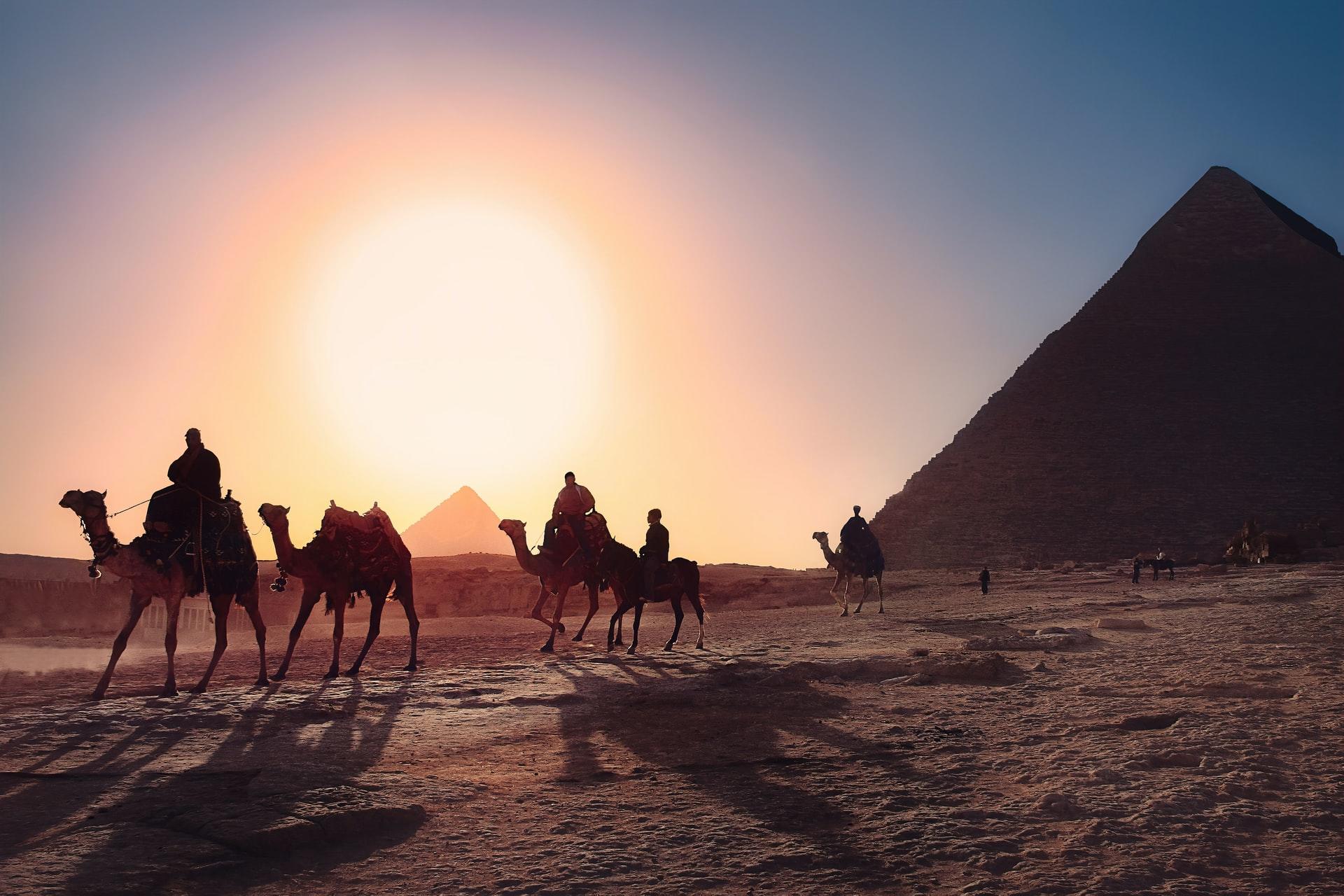 Em um deserto egípcio, sob o pôr do sol, um grupo de homens montados em camelos estão a passar.