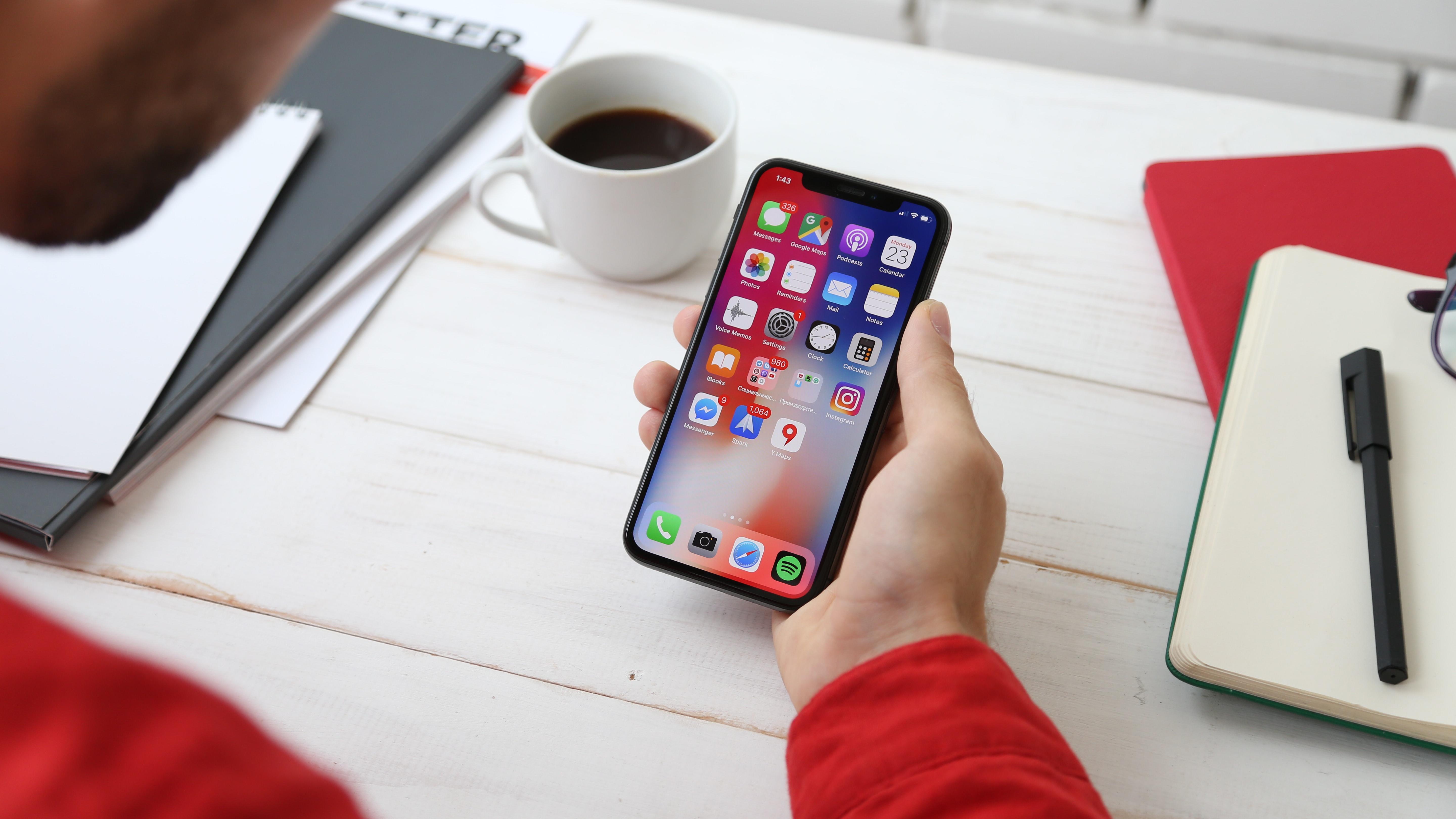 Rapaz com blusa vermelha está a segurar um celular com diversos aplicativos em destaque, tendo ao seu redor uma xícara de café, cadernos, agendas, óculos e caneta.