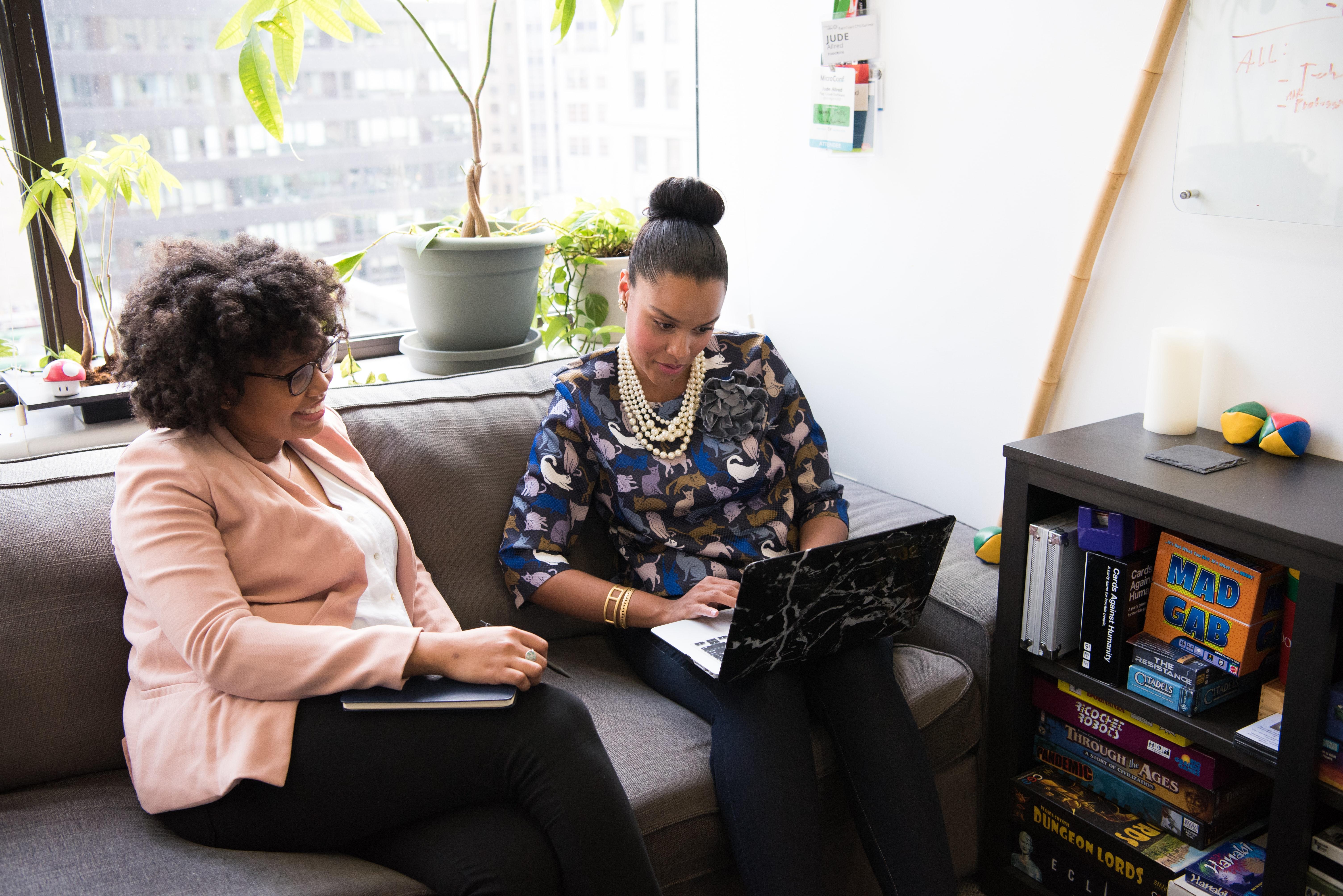 Duas mulheres em um ambiente de escritório, sendo que uma delas está a segurar um notebook aberto, estão prestando atenção no dispositivo em uma suposta comunicação.