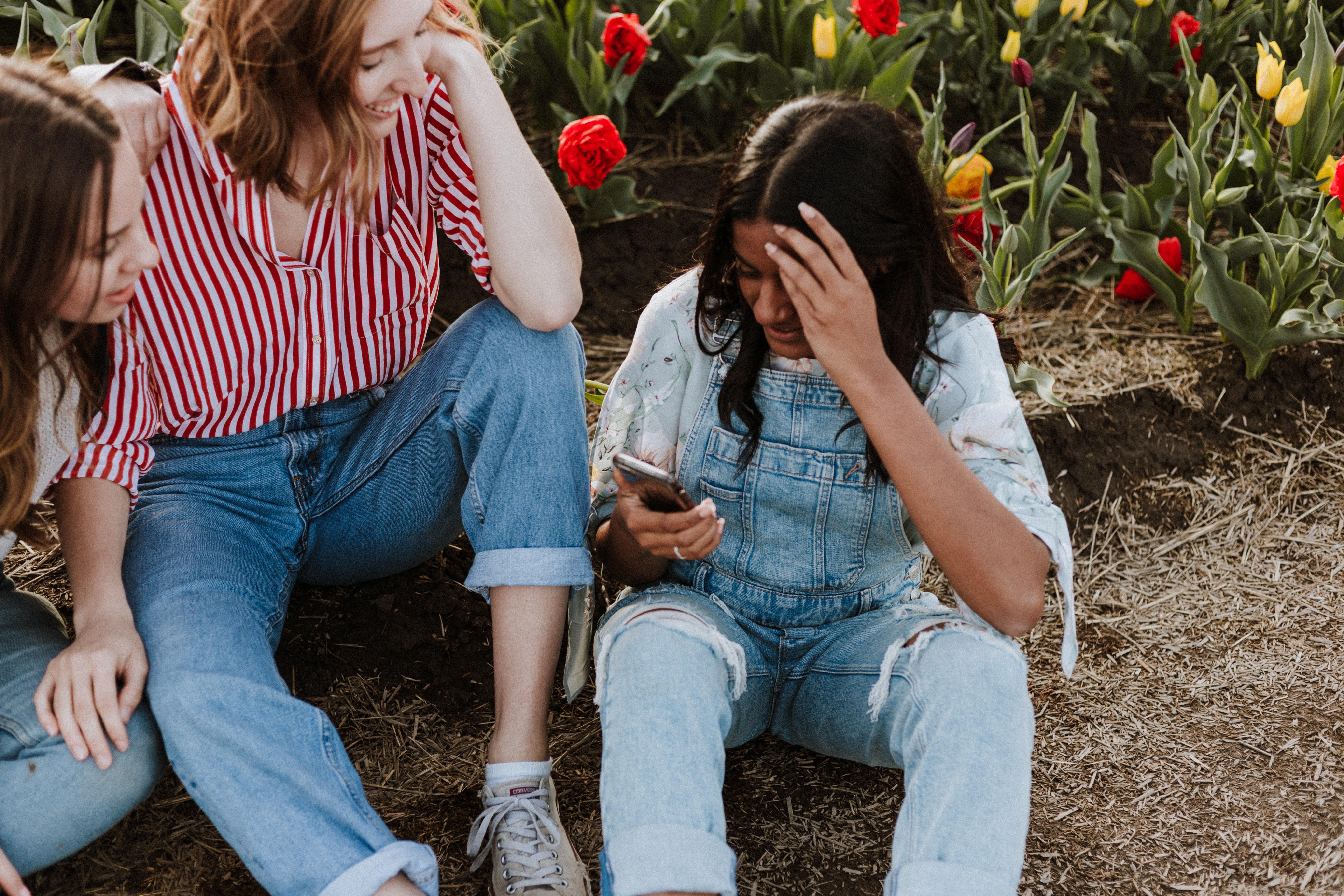 Três jovens estão sentadas em um jardim, sendo que uma está a segurar um smartphone, a atenção está voltada ao respectivo dispositivo.