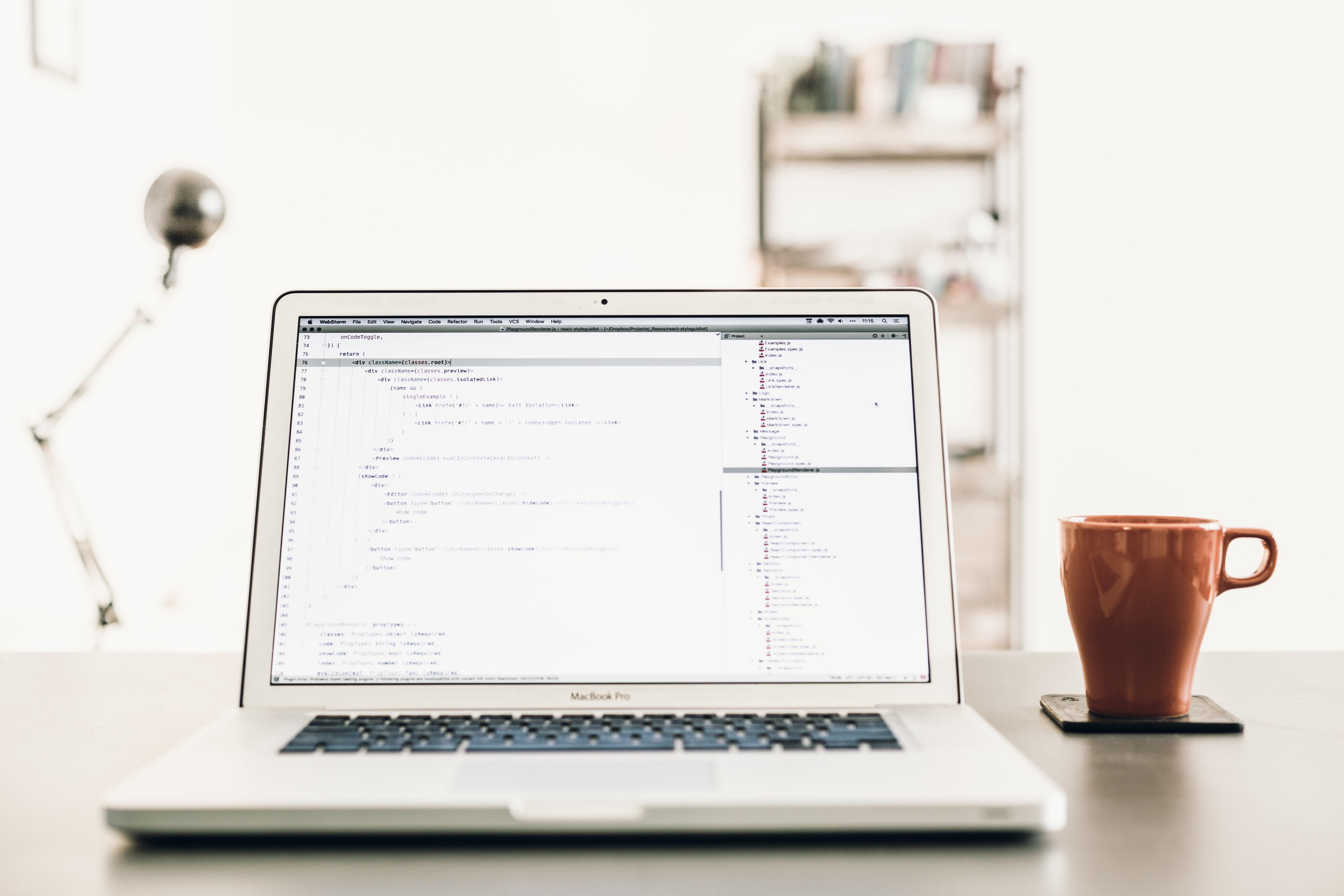 Notebook aberto tendo na tela vário códigos de Javascript enquanto que, ao lado do objeto, uma xícara vermelha.