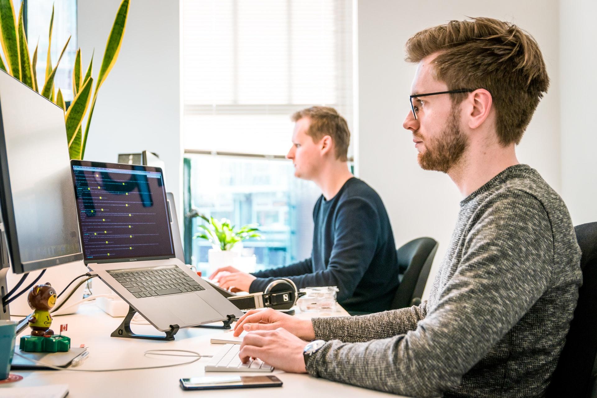 Dois rapazes estão trabalhando no escritório, concentrados em seus notebooks e desktops.
