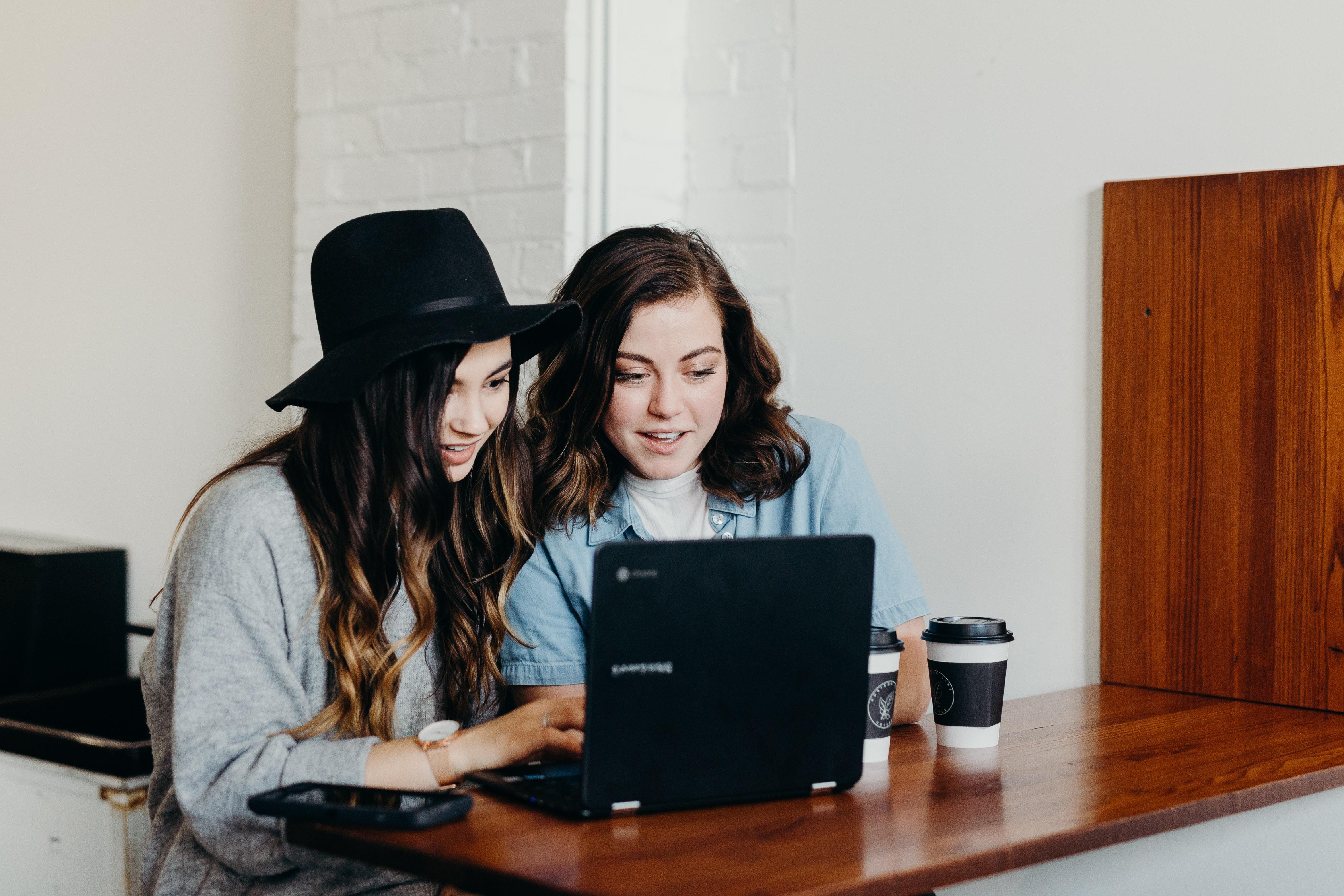 Duas jovens estão diante de um laptop aberto e se mostram atentas na comunicação.