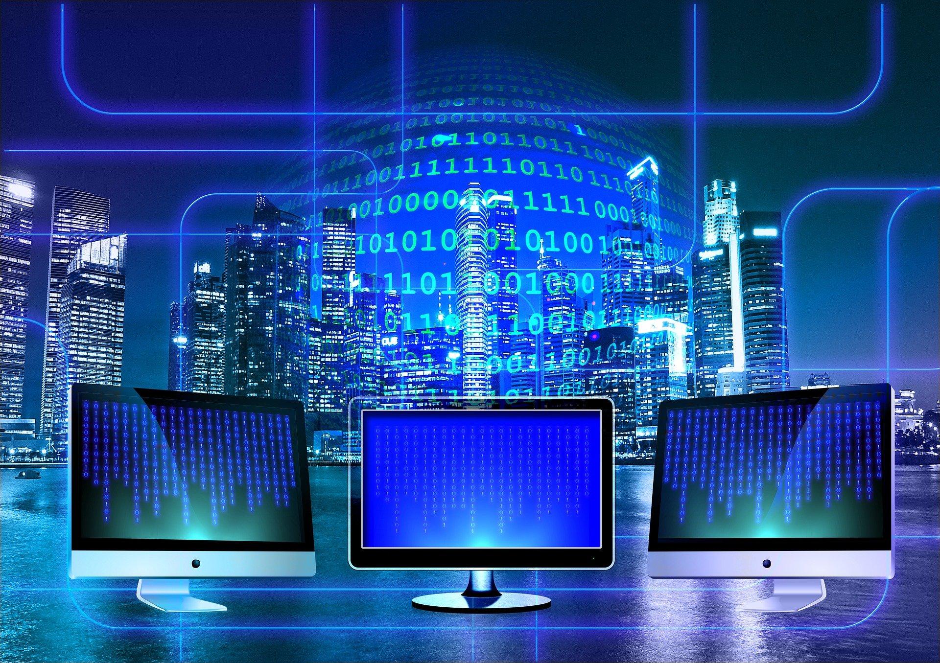 Três monitores de destacam junto com códigos preenchendo toda a imagem e, ao fundo, uma metrópole toda iluminada à noite.