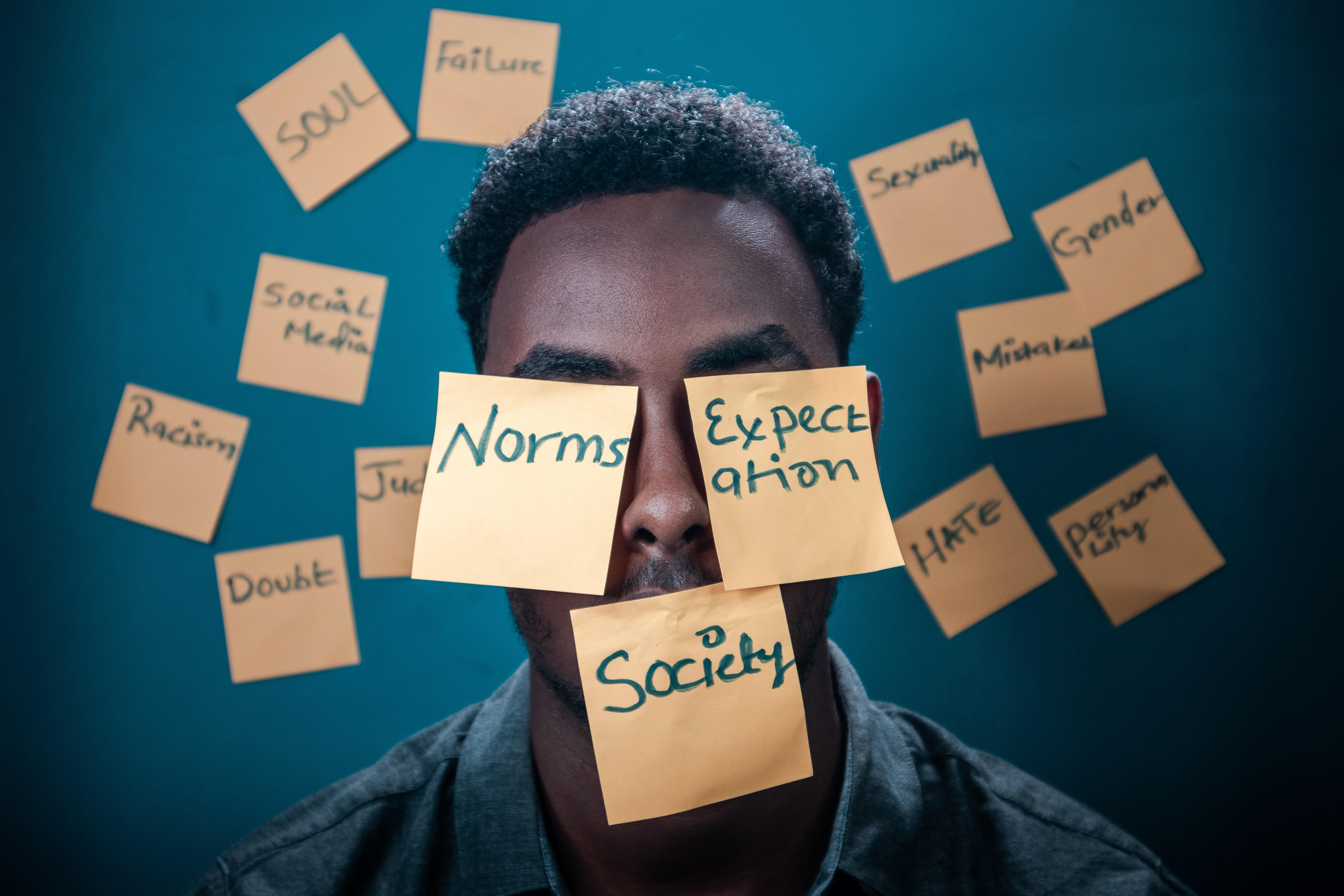 Imagem de um rapaz tendo no rosto três post its colocados com as seguintes palavras: "Norms", "Expectation", Society". Atrás dele também há outros post its colados com outras palavras.