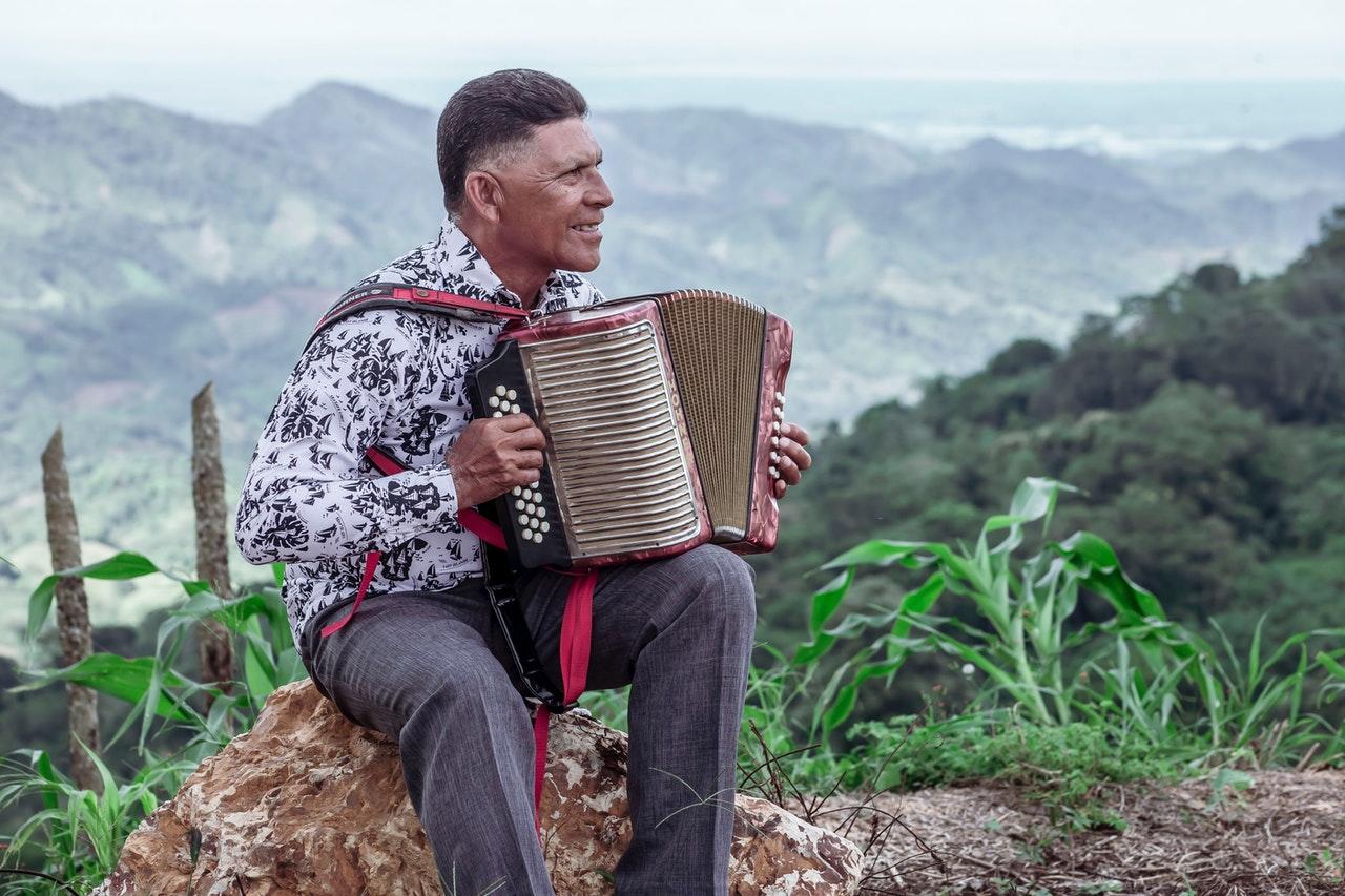 Homem sentado em uma pedra está sorrindo tocando acordeon, tendo atrás dele uma bela paisagem da natureza.