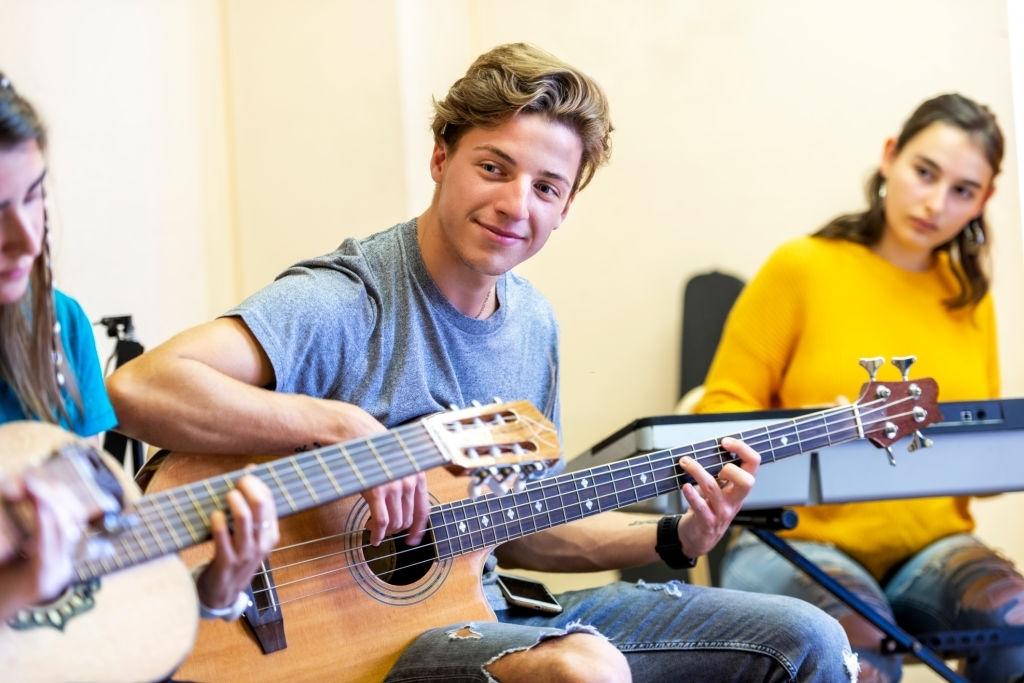 Em uma sala de estudo musical, rapaz está aprendendo a tocar contrabaixo ao lado de uma moça tocando violão e outra no teclado.
