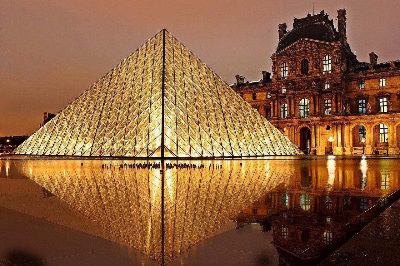 Vista externa noturna do Museu do Louvre e sua icônica pirâmide de vidro iluminada.