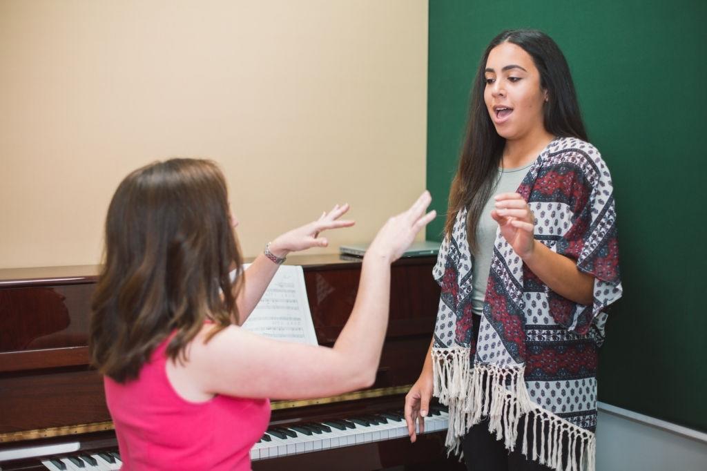 Professora de canto, ao lado de um piano, está a ensinar uma jovem aluna técnicas de respiração, postura e dicção.