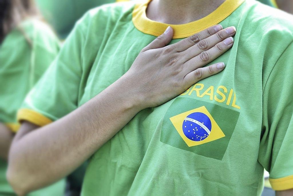 Pessoa vestida com uma camiseta verde e amarela com a mão sobre o peito e, tendo ao lado, como destaque, a bandeira do Brasil e, além disso, escrito logo acima a palavra “Brasil”.
