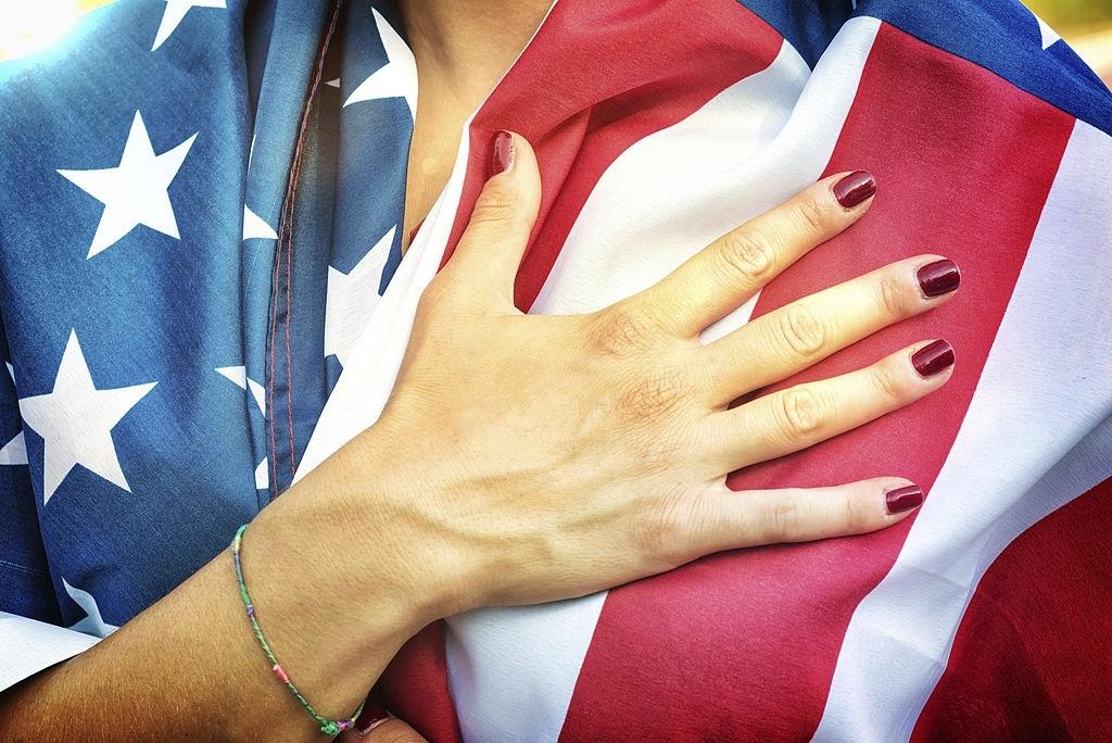 Mulher com a bandeira americano sobre o corpo está com a mão direita na altura do peito para entoar o Hino Nacional dos Estados Unidos.