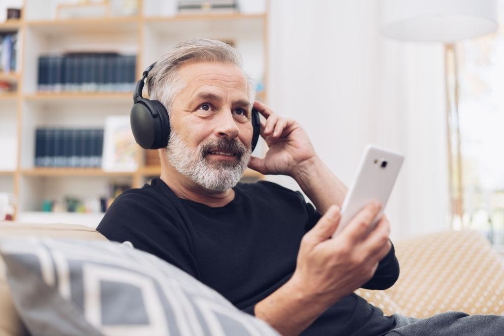Homem de meia idade, em casa, escutando o hino pelo fone de ouvido o qual está conectado via bluetooth em um smartphone.