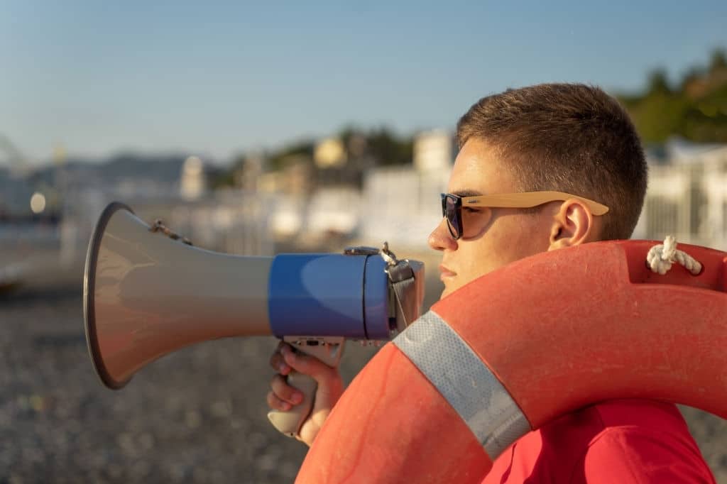 Salva-vidas usando óculos escuros está com um megafone e boia, alertando pessoas na praia.