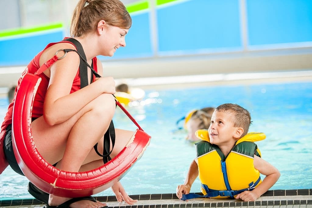 Mulher salva-vidas conversando com um menino que está usando um colete salva-vidas na piscina durante uma aula de natação.