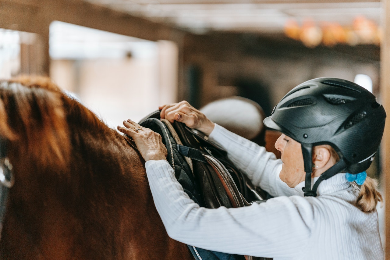 Mulher idosa usa capacete e segura cela posicionada no cavalo. Ela parece que está pronta para montar no animal.
