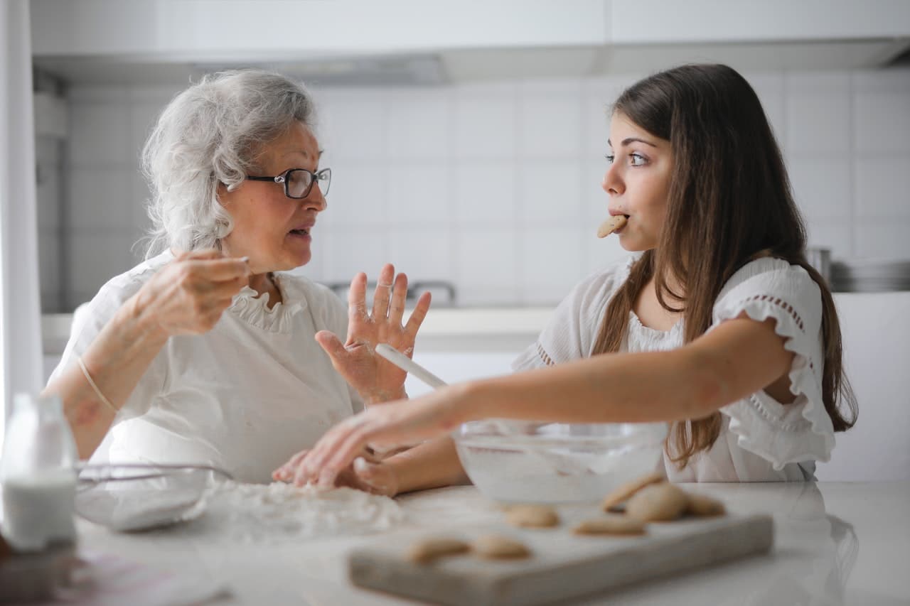 Idosa e adolescente, provavelmente avó e neta, fazem receita de biscoito juntas na cozinha.