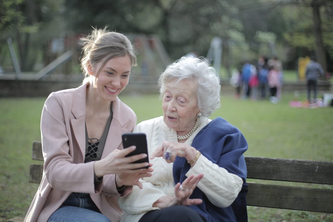 Sentadas no banco de um parque, jovem mulher mostra telefone celular para mulher idosa, que possui expressão de surpresa no rosto.