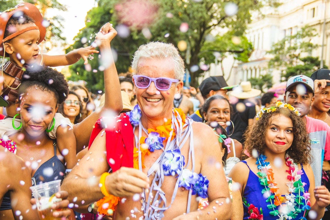 Homem de cabelos brancos usa óculos de lente azul e colar de flores. Ele está no meio da multidão em um bloco de carnaval.