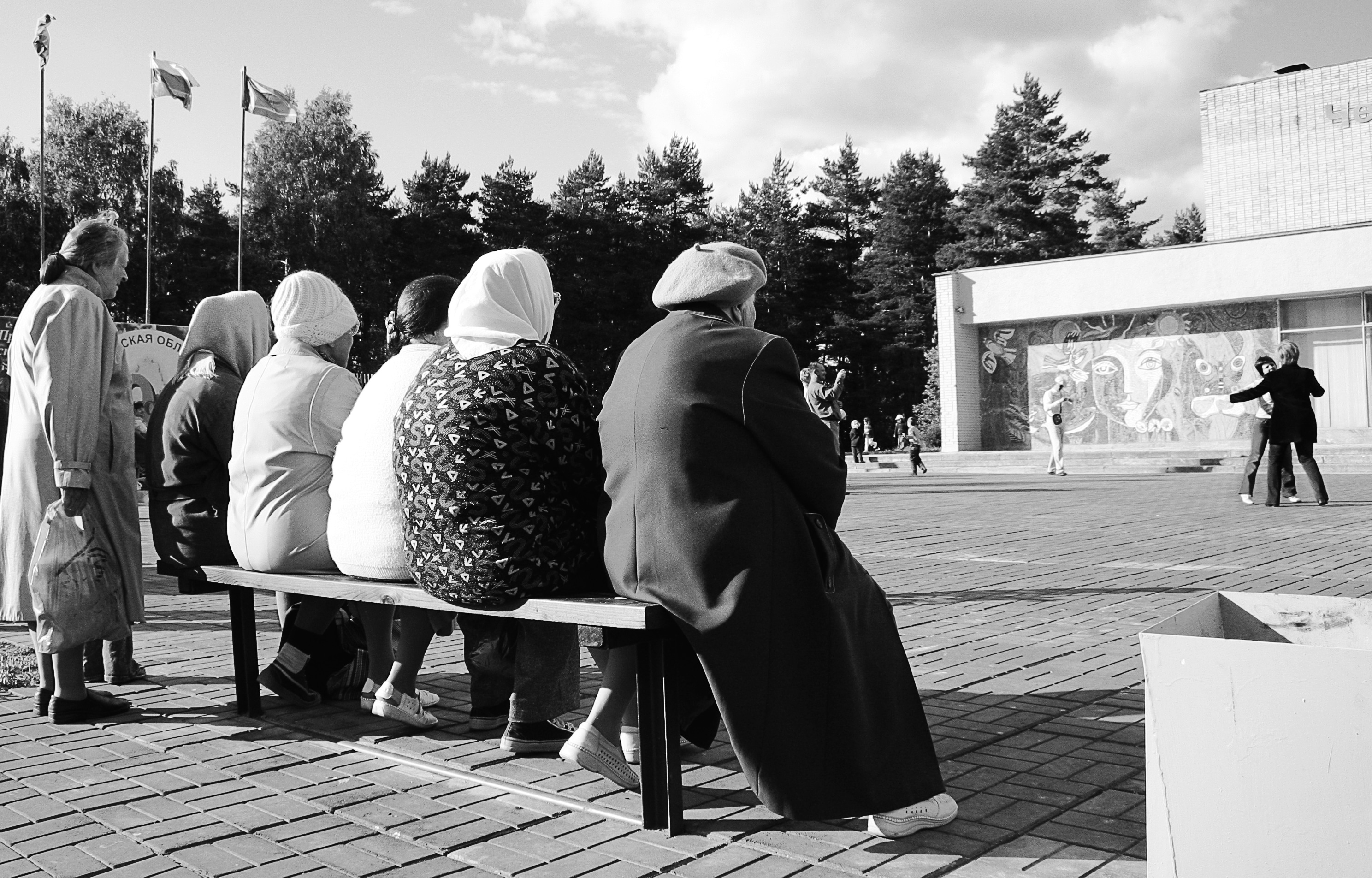 Cinco idosos sentados lado a lado em um banco de praça observam um casal dançando. Um outro idoso também observa em pé atrás dos cinco sentados.