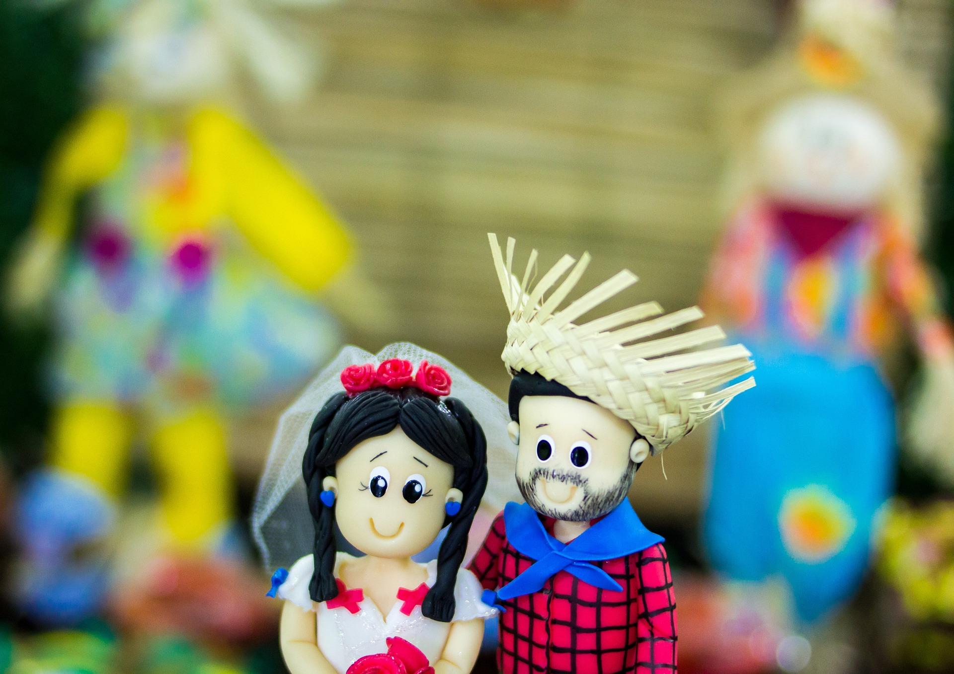 Fundo desfocado. No destaque da imagem, é possível ver um casal de bonecos de biscuit caracterizado de motivos juninos. 