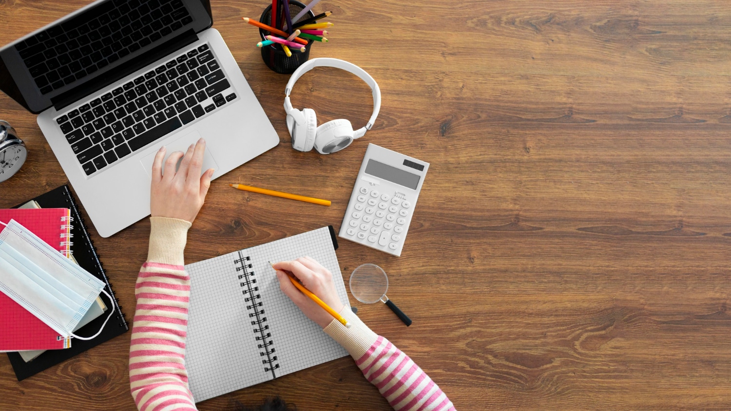 Mãos usando pc e caderno para anotar informações, posados em uma mesa.