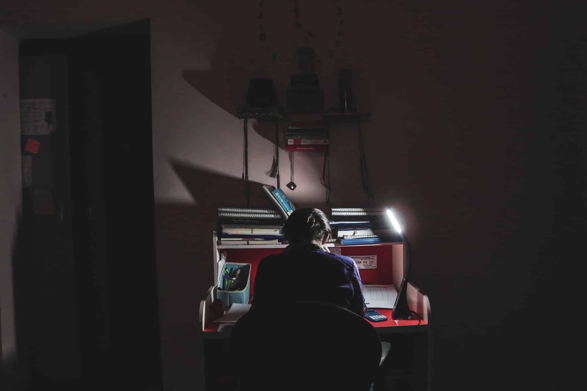 Estudante sentado em escrivaninha em uma sala escura estudando.