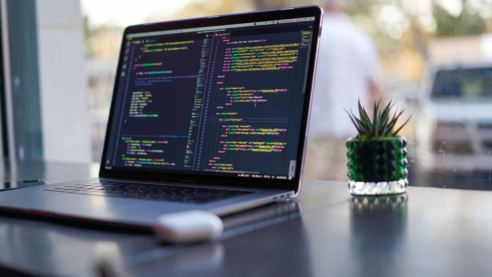 Computador com códigos exibidos na tela. Ao lado um vaso decorativo de suculenta.