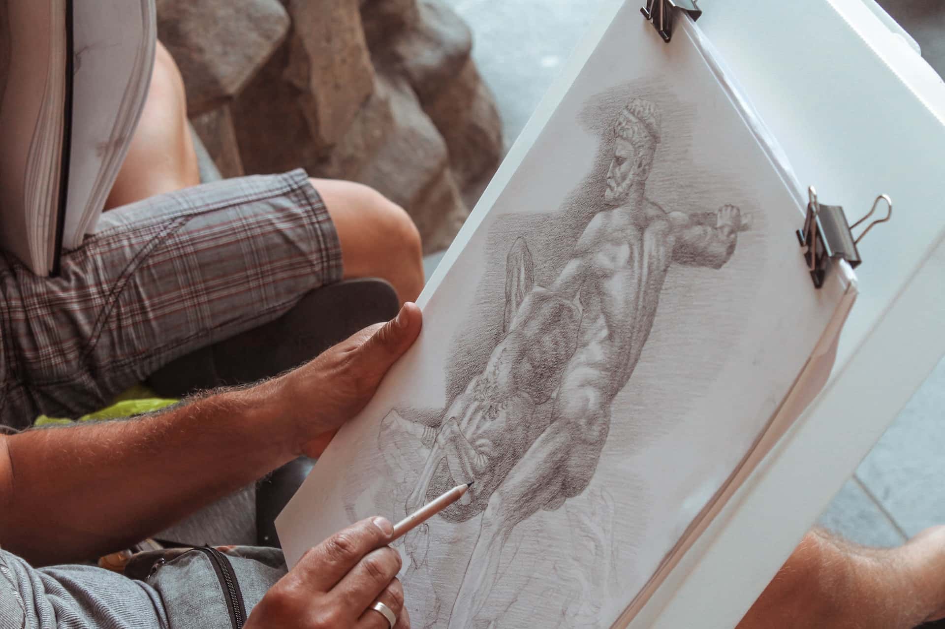 Homem reproduzindo em um desenho  uma estátua à lápis em um papel.