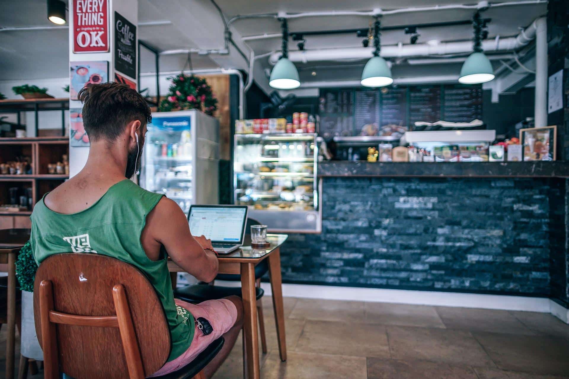 Homem de bermuda, camiseta e fones no ouvido trabalhando em um computador portátil em um café.