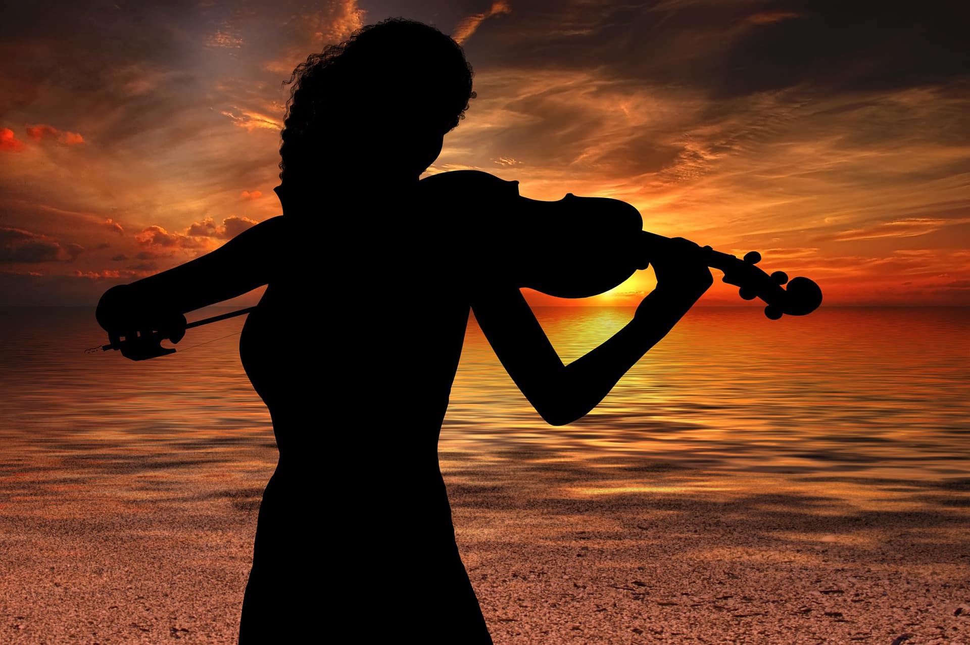 Sobra de uma mulher tocando violino em uma praia com o sol se pondo ao fundo.
