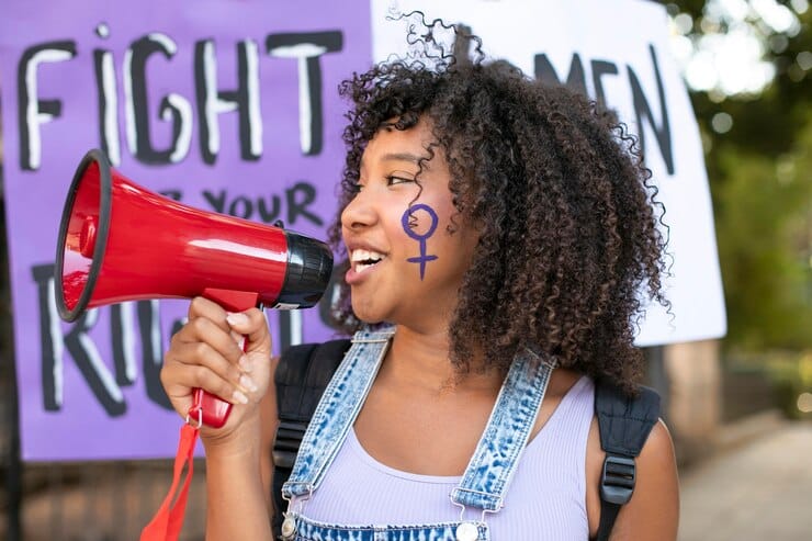 Mulher fala ao microfone em busca de direitos iguais. Seu rosto carrega a marca do feminismo.